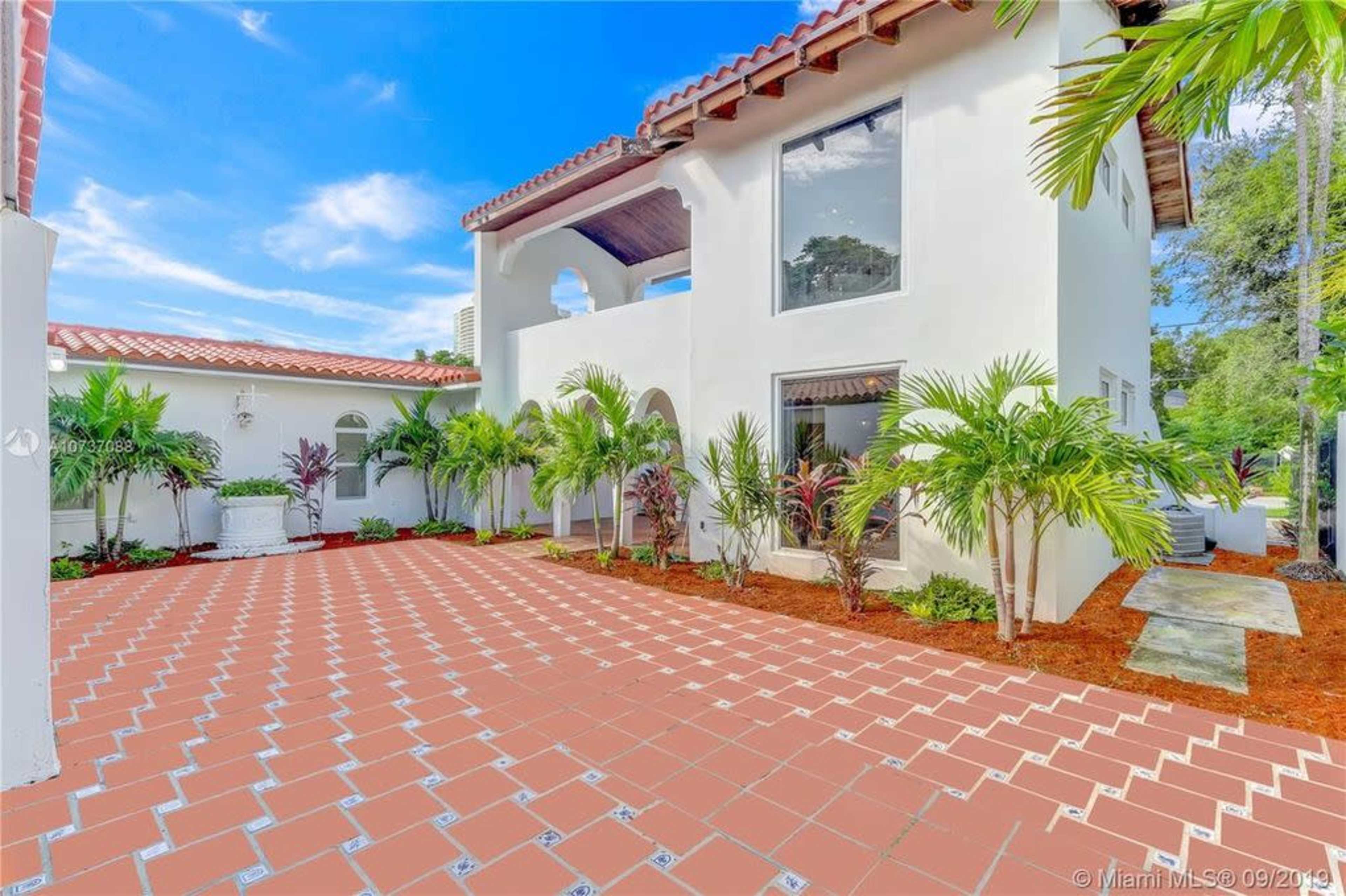 A two-story white house with a red tile roof surrounded by tropical plants and a patterned terracotta patio.