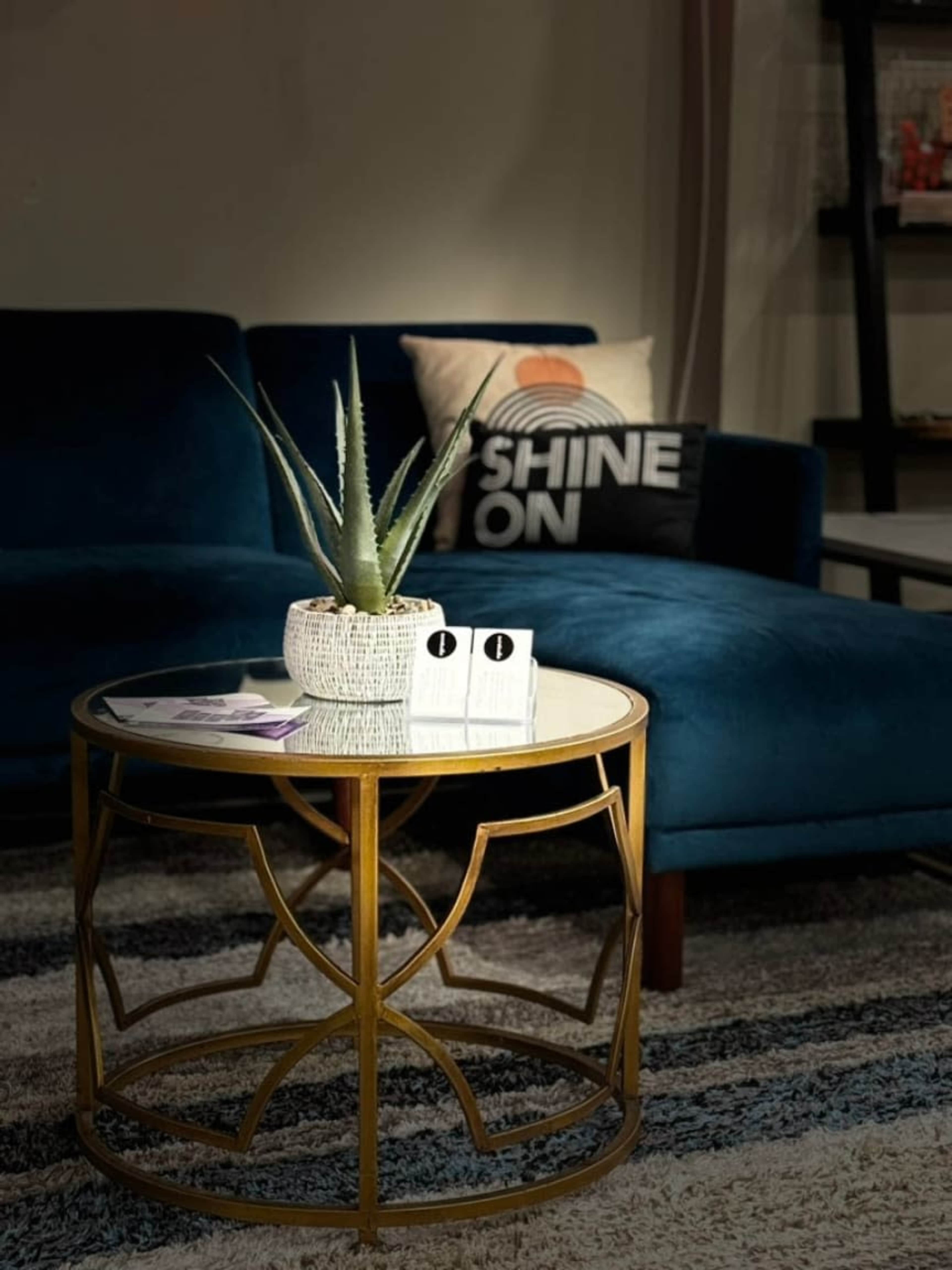A round gold coffee table with a potted plant sits in front of a blue sofa, accompanied by a decorative pillow and magazines on the table.