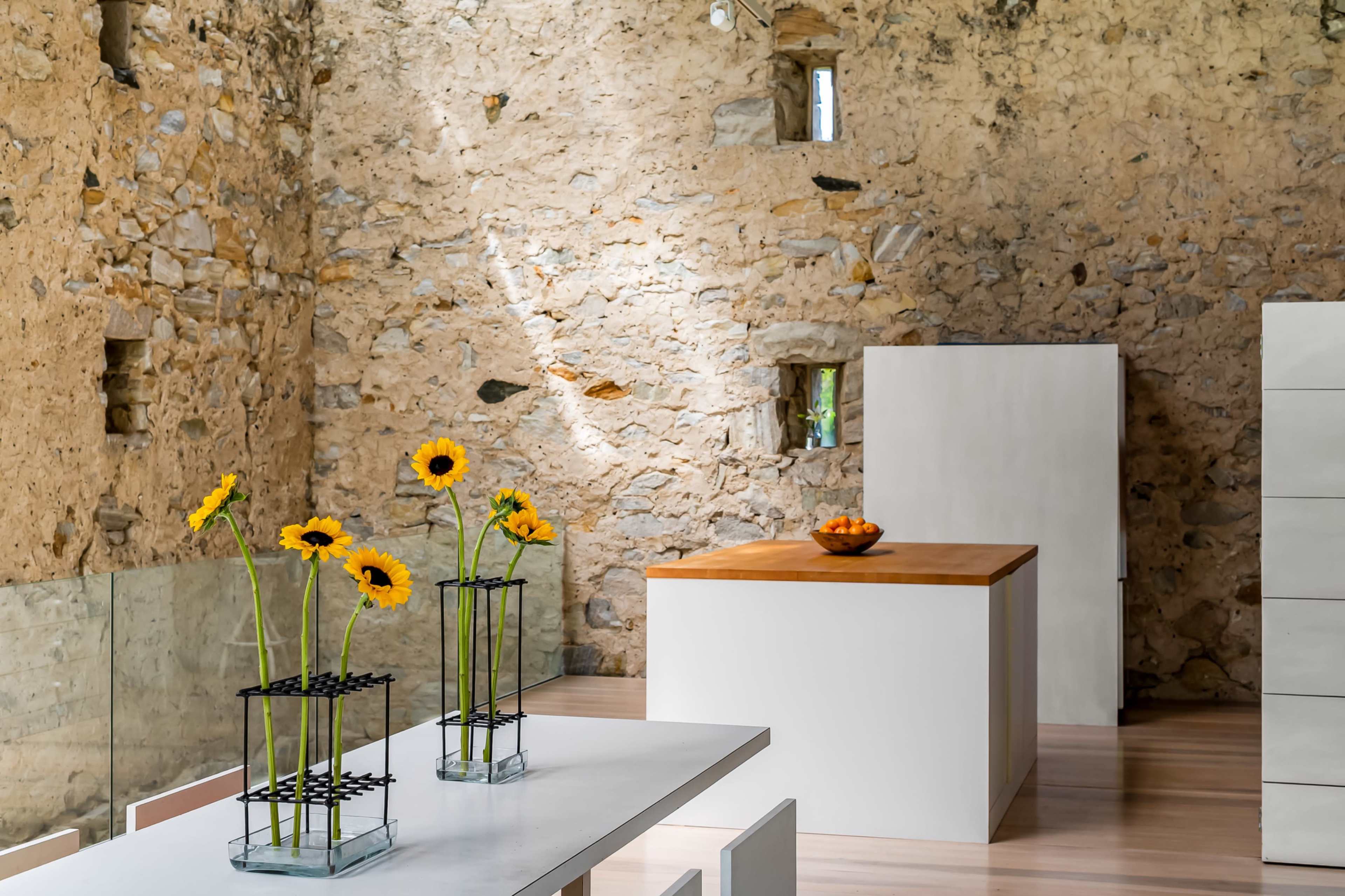 The image depicts a modern kitchen interior with stone walls, a wooden island, and vases of sunflowers on a white dining table.