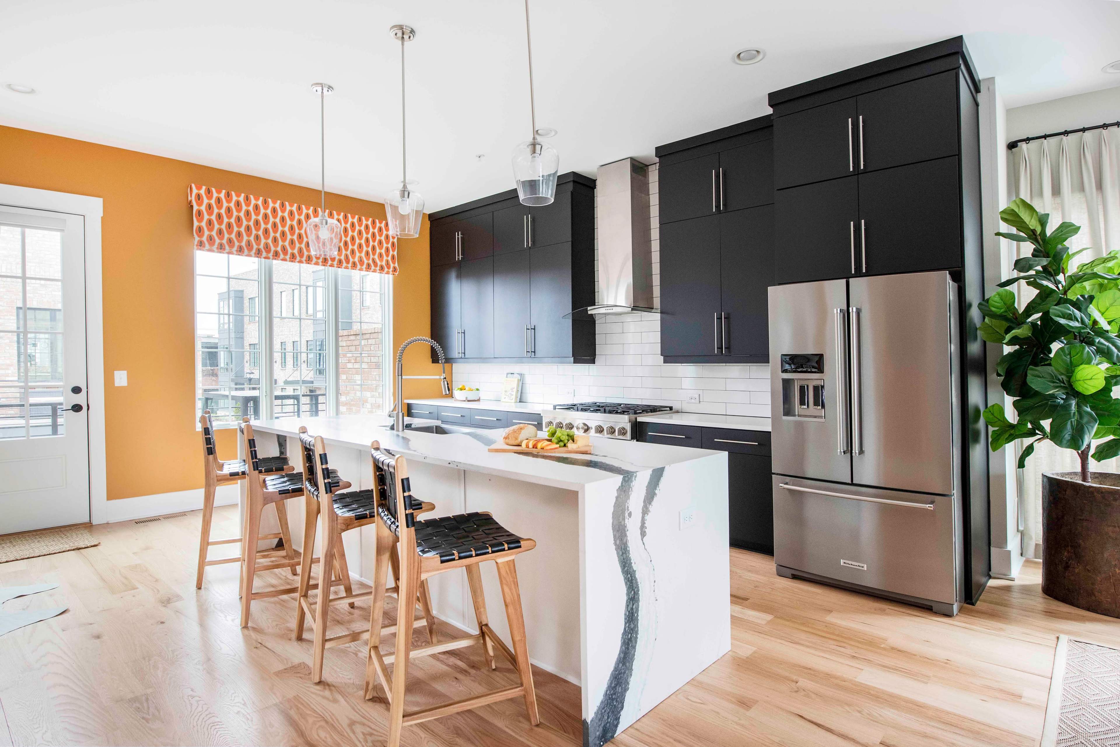 A modern kitchen features black cabinetry, a stainless steel refrigerator, and a central island with bar stools, illuminated by a pendant light.