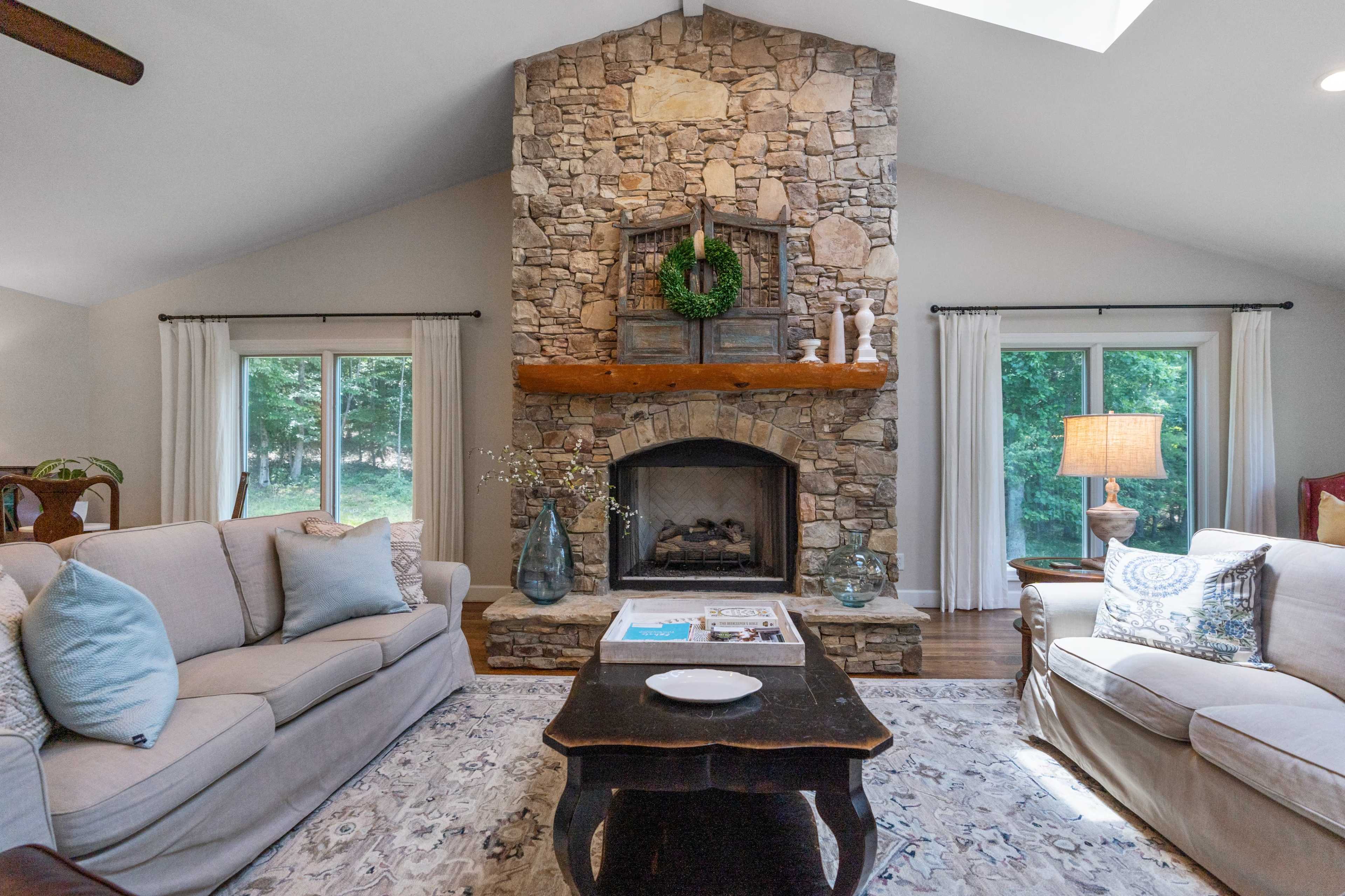 The living room features a large stone fireplace with a wooden mantel, surrounded by two sofas and natural light from large windows.