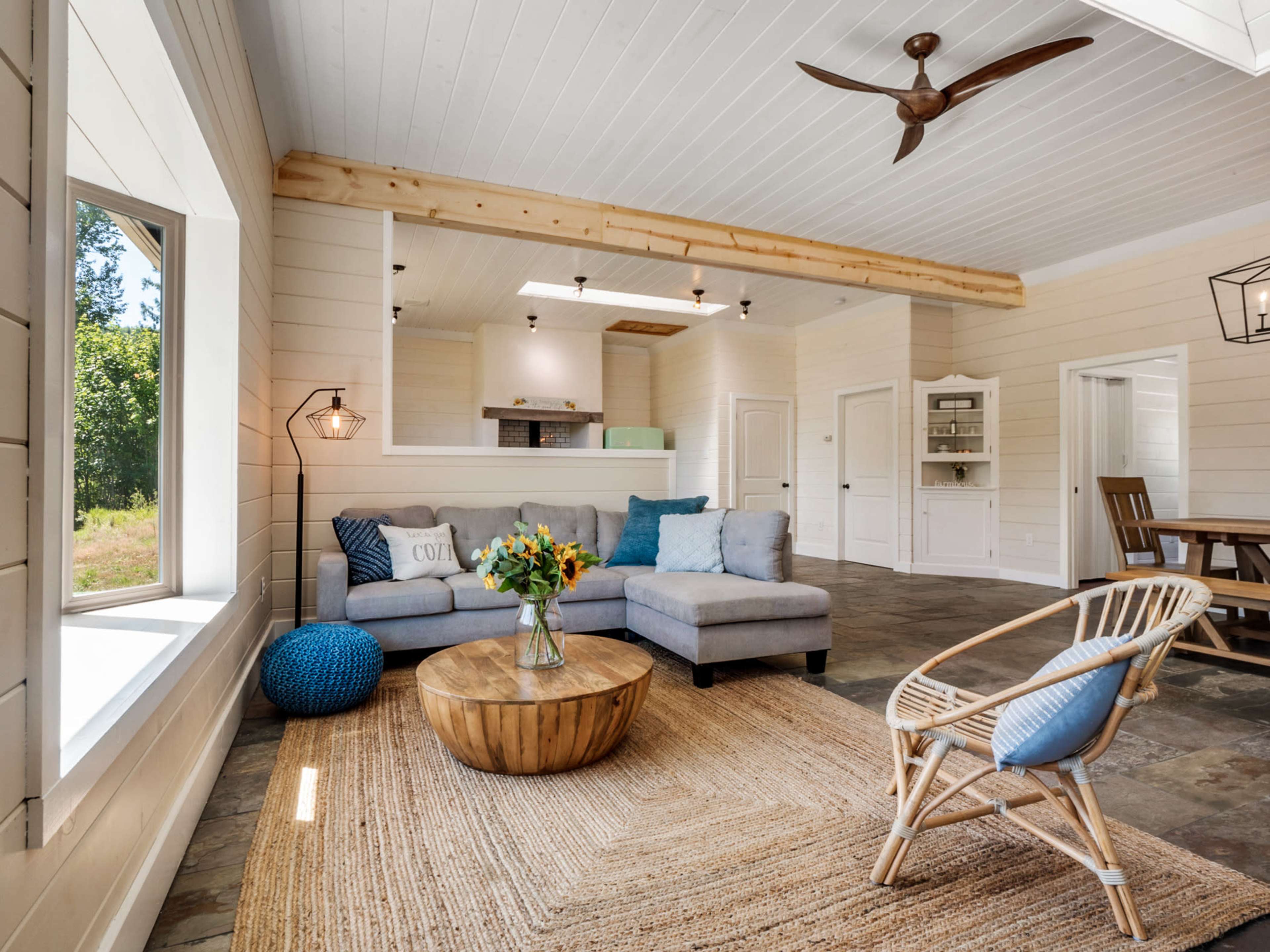 The image shows a bright, open living space featuring a gray sectional sofa, a round wooden coffee table, and a rattan chair, surrounded by light-colored walls and natural elements.