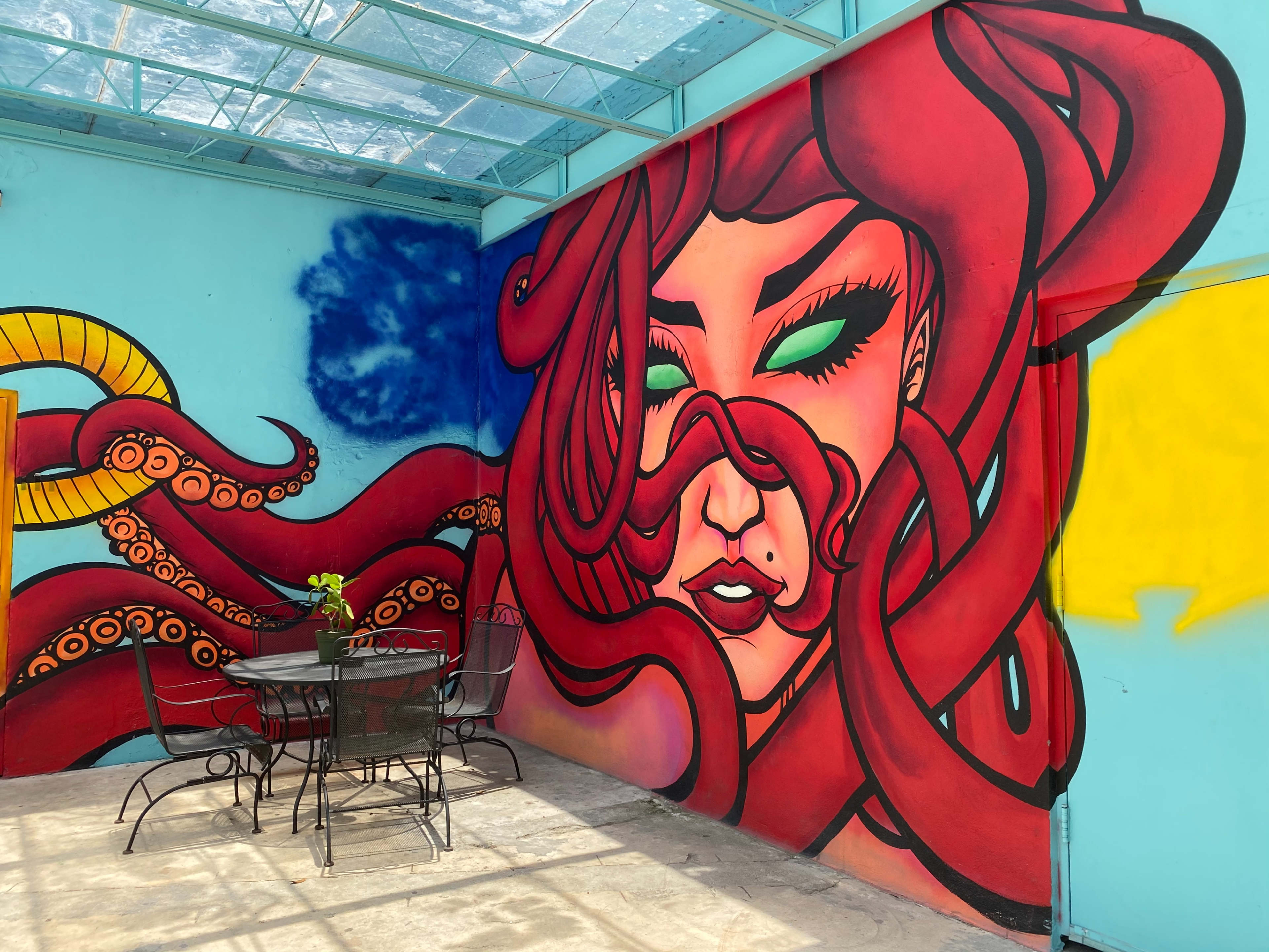 The image shows a vibrant mural featuring a red-haired woman intertwined with abstract shapes, set against a blue and yellow background, alongside a small black table with four chairs in a bright room.