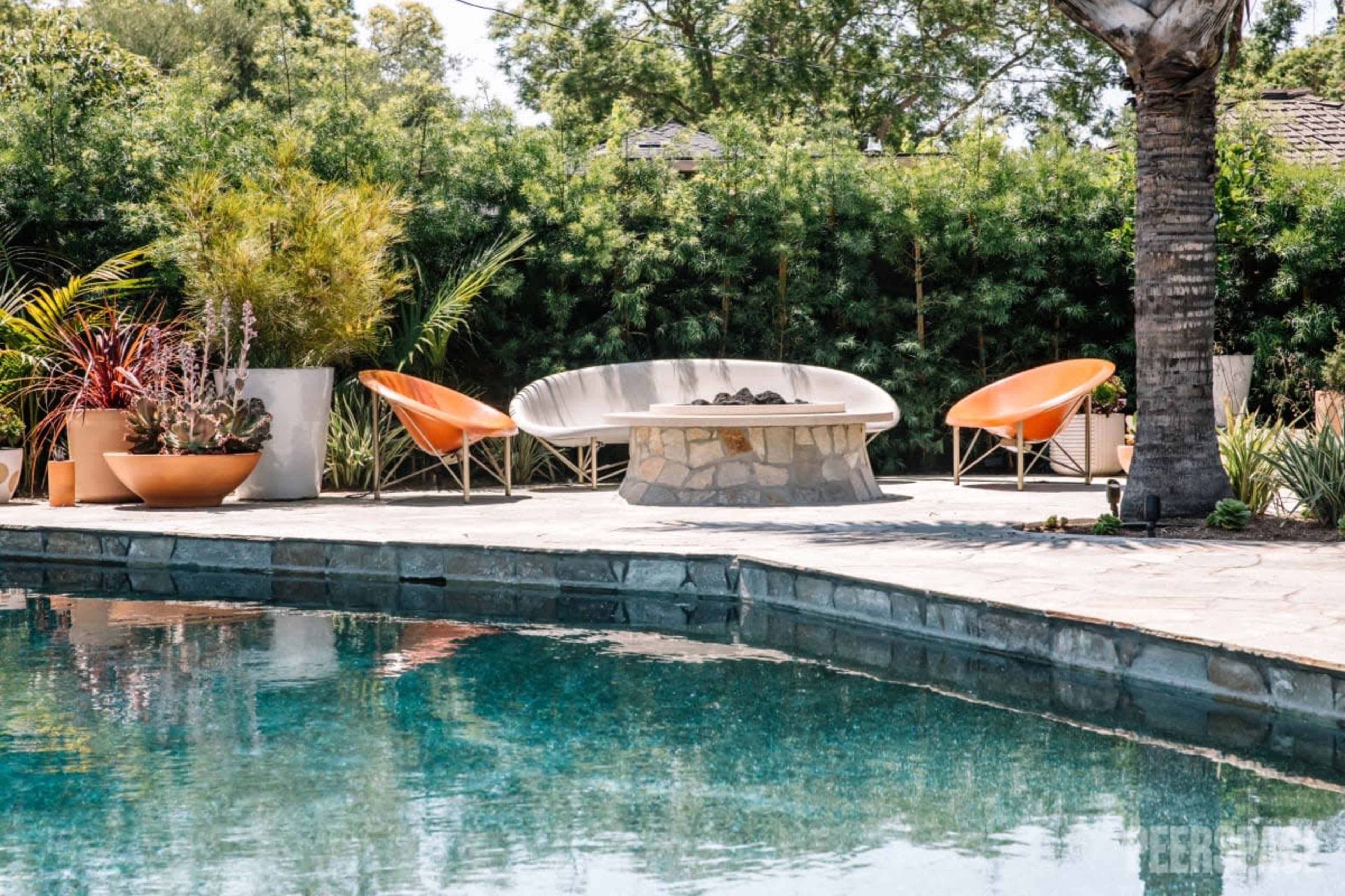 A modern outdoor seating area features orange chairs and a stone fire pit beside a swimming pool surrounded by greenery.