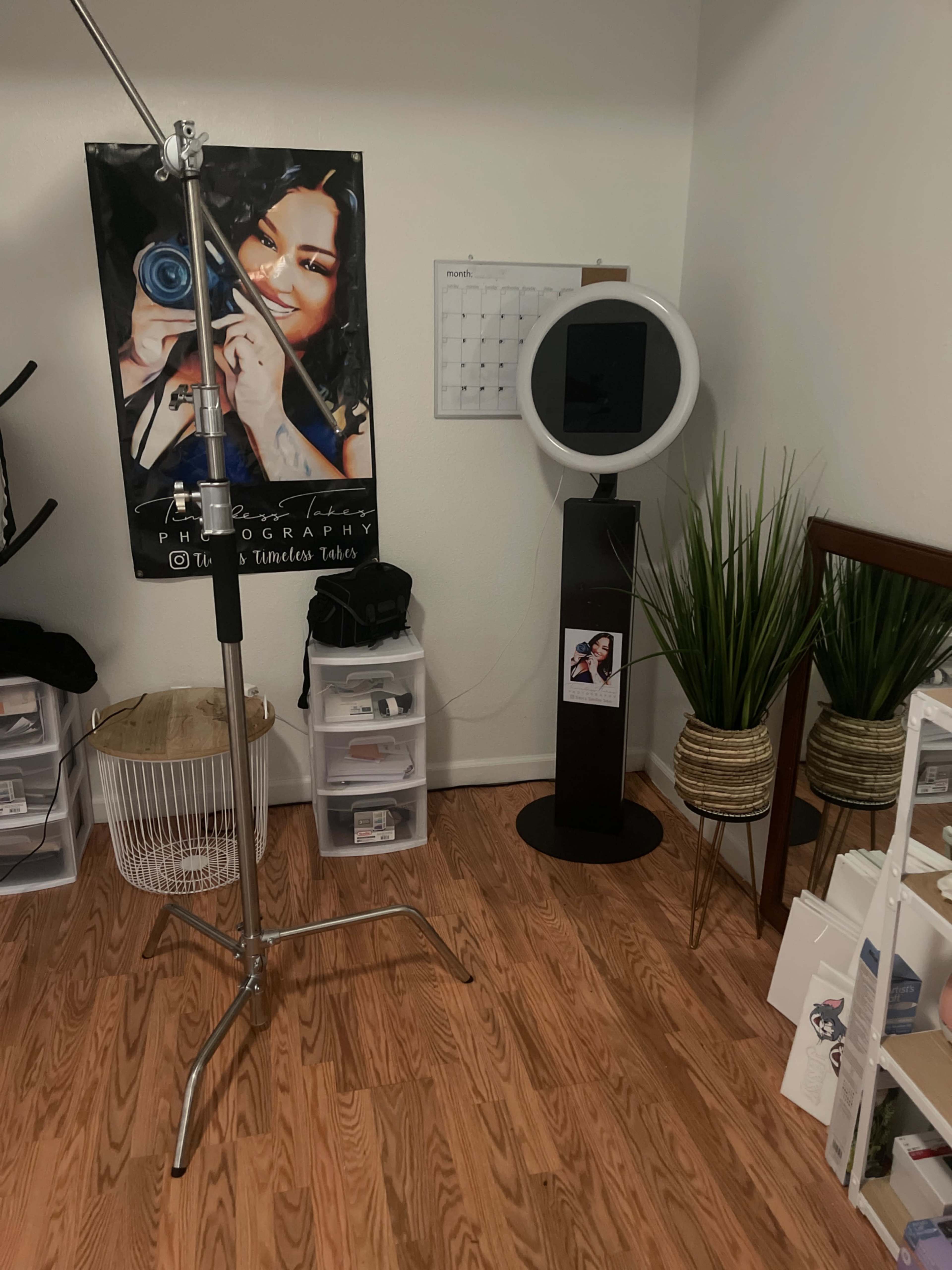 A photography studio with a backdrop featuring a woman's portrait, a photo booth setup, and organized storage containers along with decorative plants.