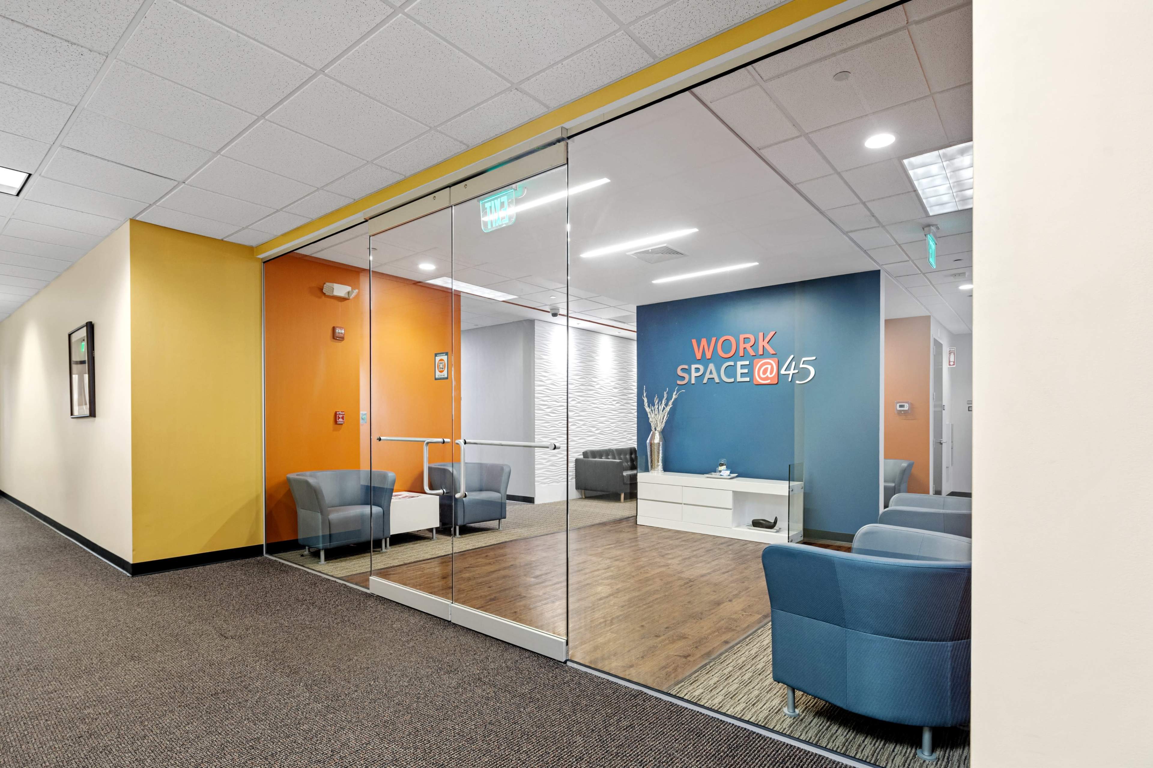 A modern office space with a glass entrance, featuring blue seating, a white table, and a wall with the text "WORK SPACE @ 45" in orange.