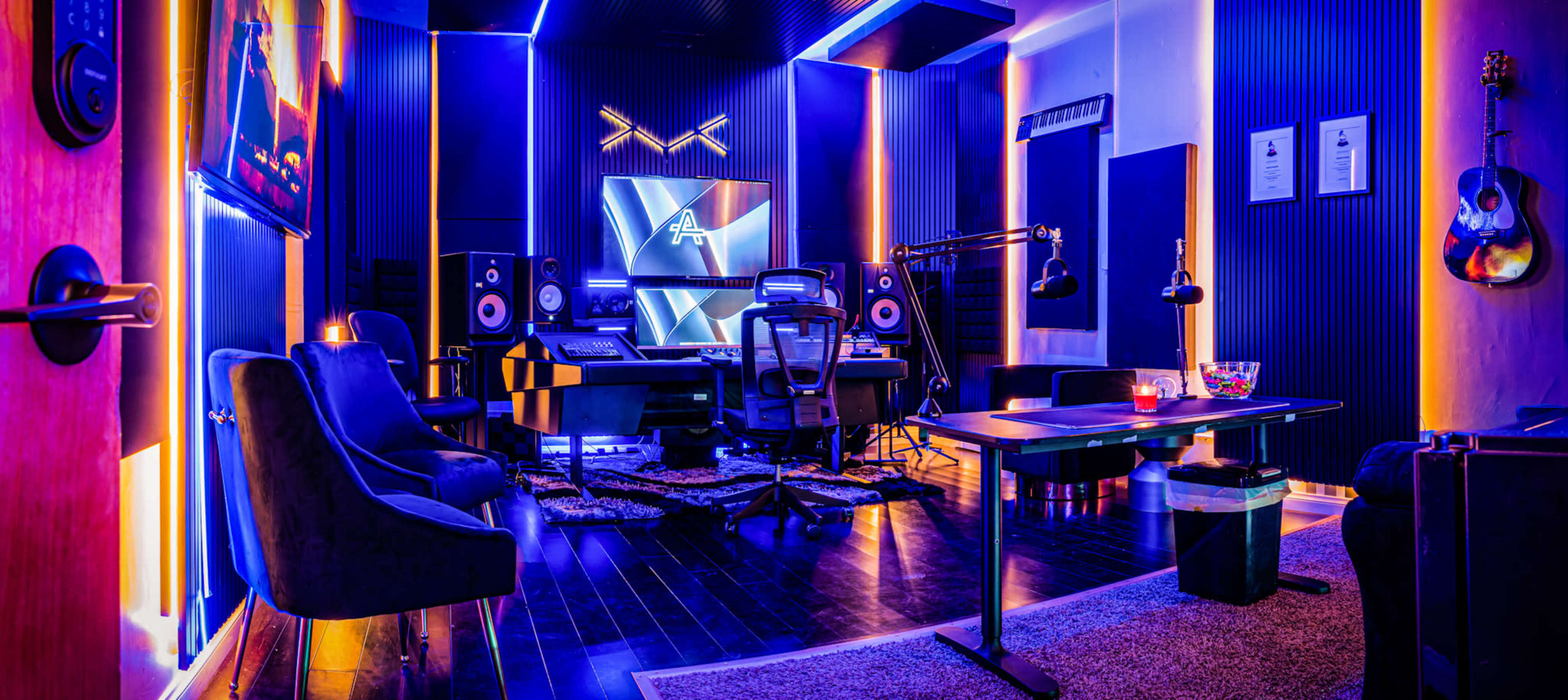 A modern recording studio features a mixing desk, speakers, and colorful LED lighting, with seating and acoustic treatment on the walls.