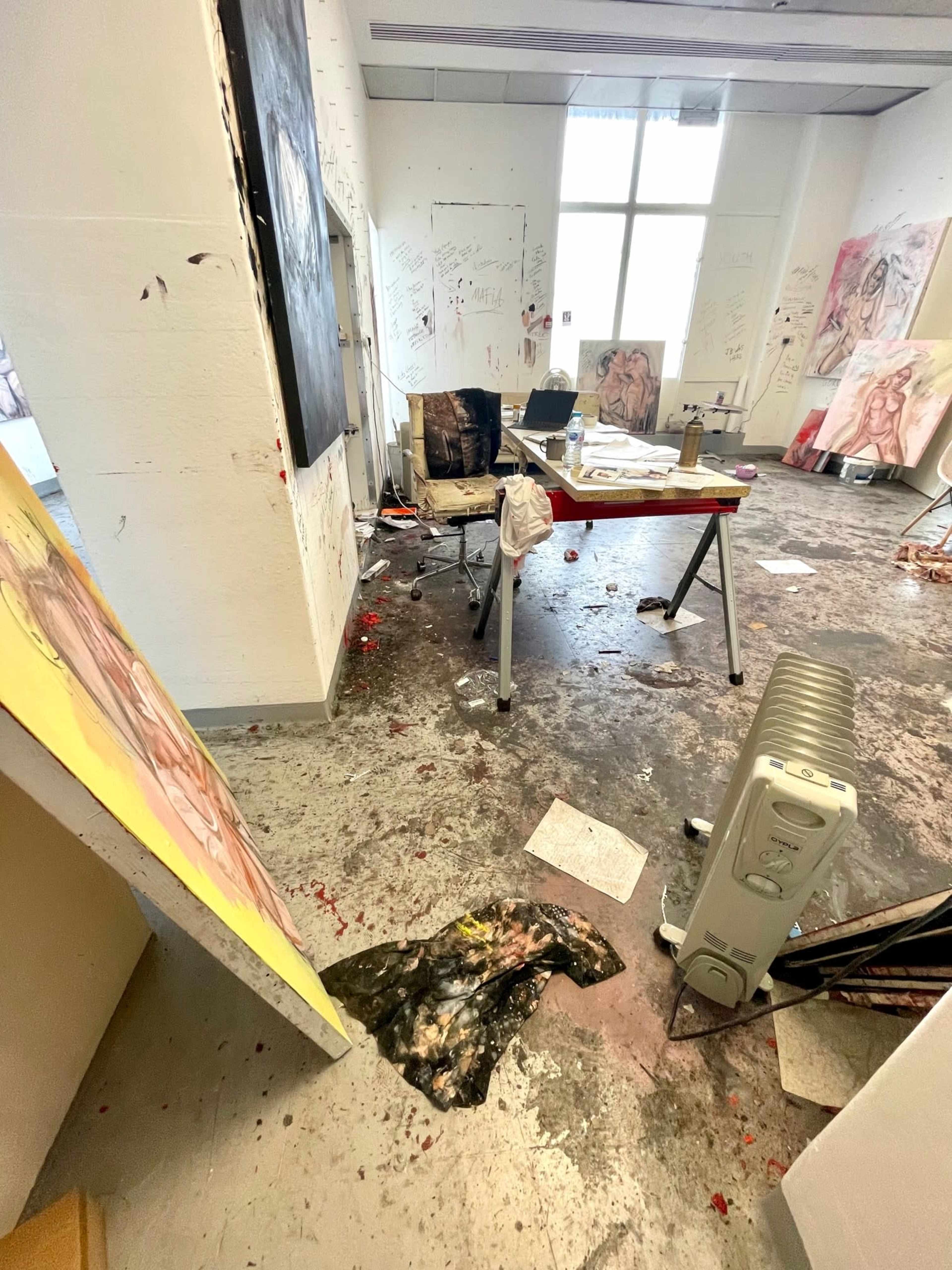 A cluttered artist's studio with scattered paintings, paint spills on the floor, and disorganized furniture.
