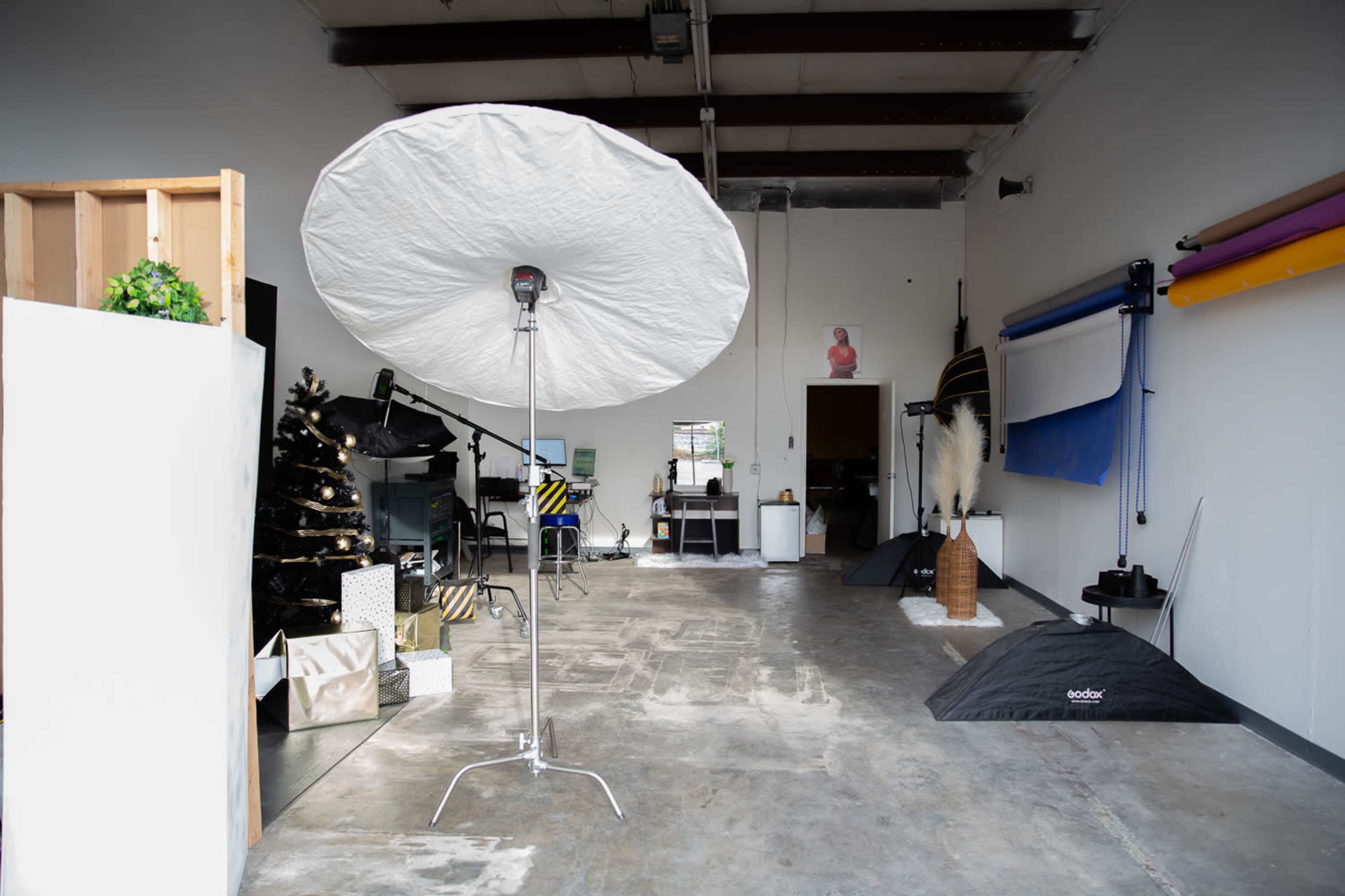 The image depicts a photography studio with lighting equipment, backdrops, and props arranged in a spacious, minimally decorated area.
