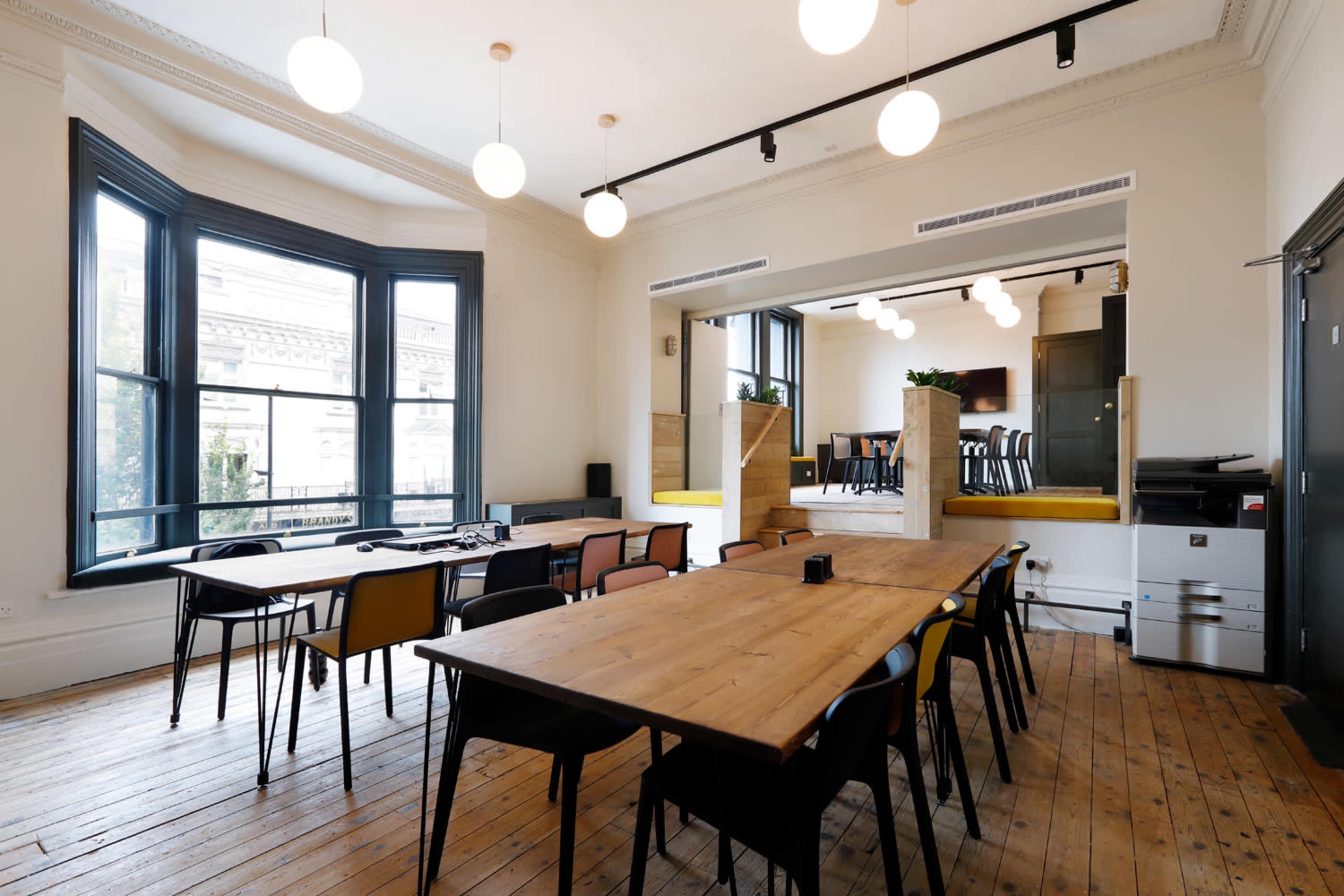A modern, well-lit meeting room features wooden tables and multi-colored chairs arranged for collaboration, with large windows providing ample natural light.