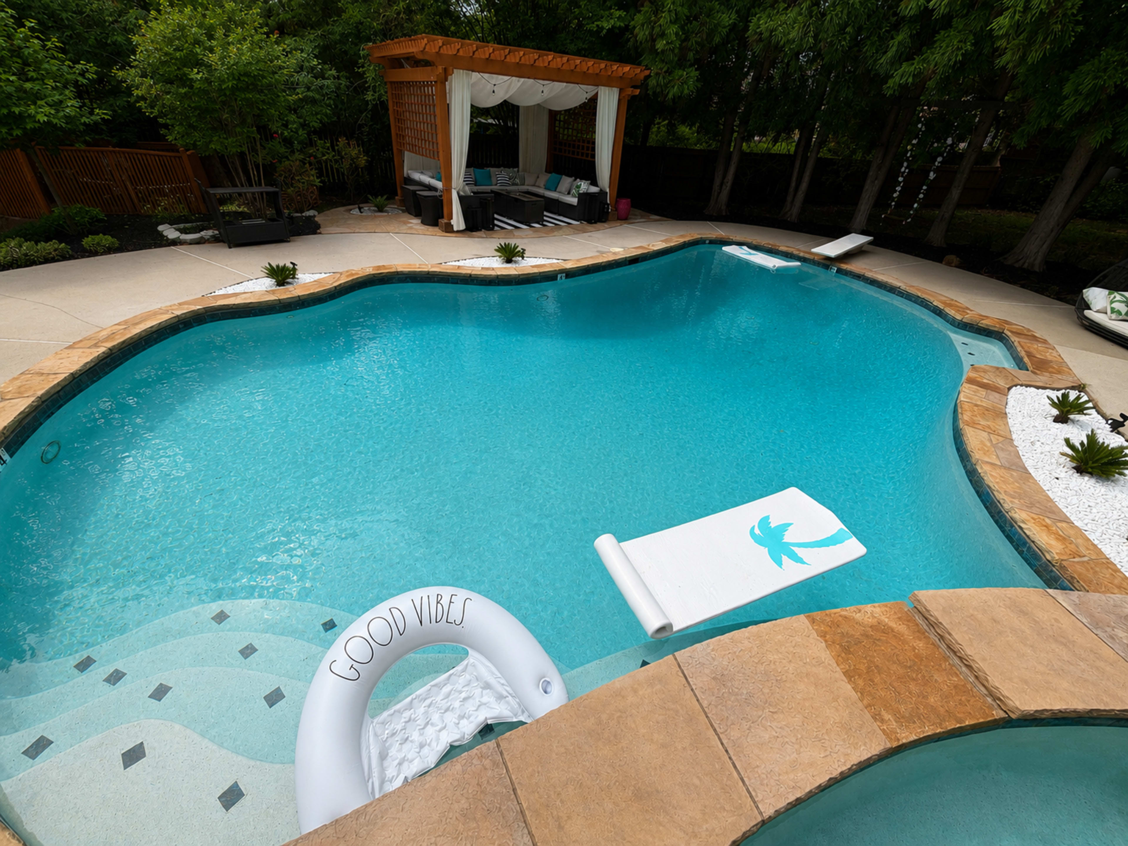 A swimming pool with a "Good Vibes" float, surrounded by landscaped greenery and a shaded seating area.