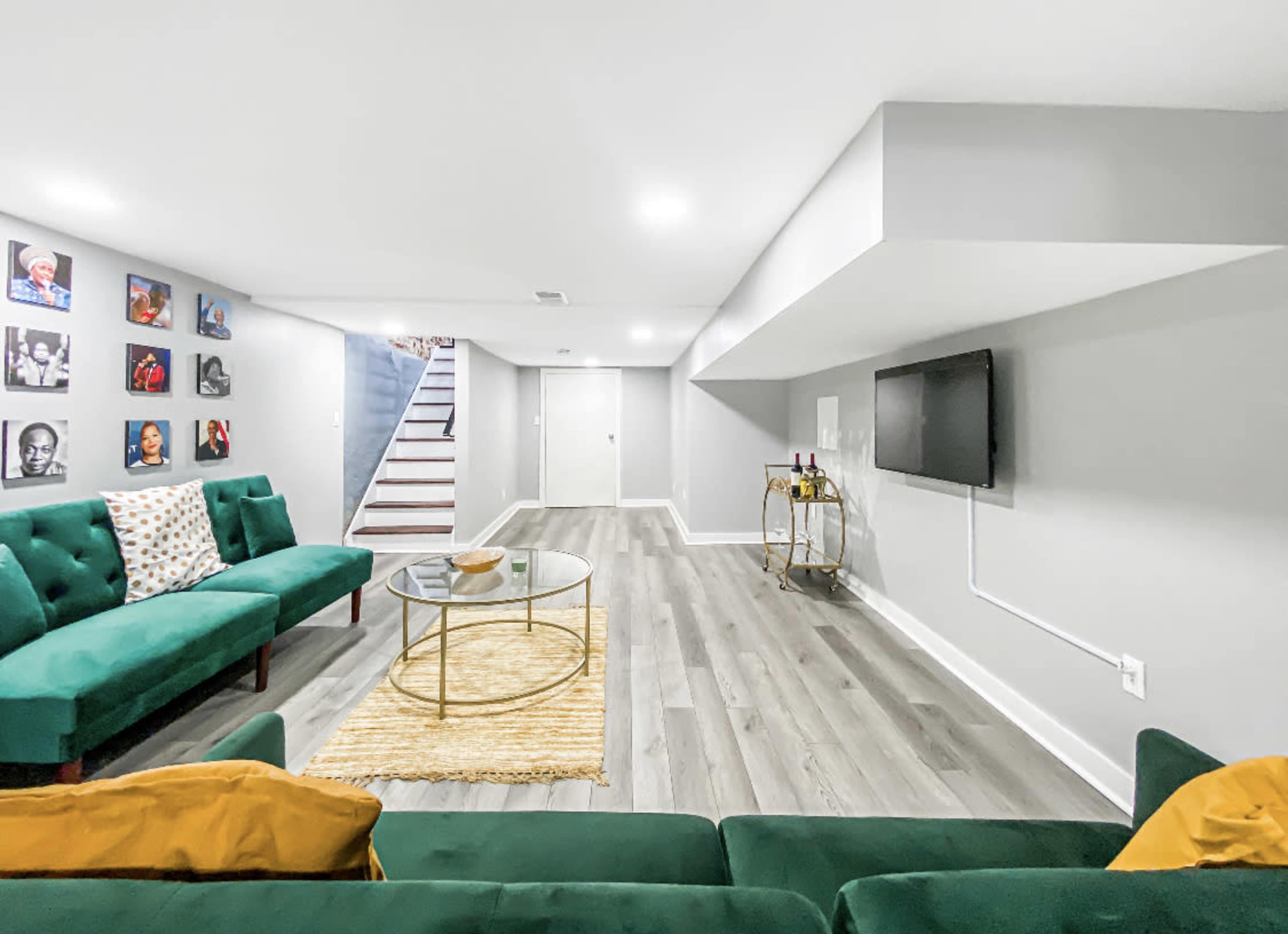 The image shows a modern basement living area featuring a green sofa, a round glass coffee table, a media console with a wall-mounted TV, and framed portraits on the walls.