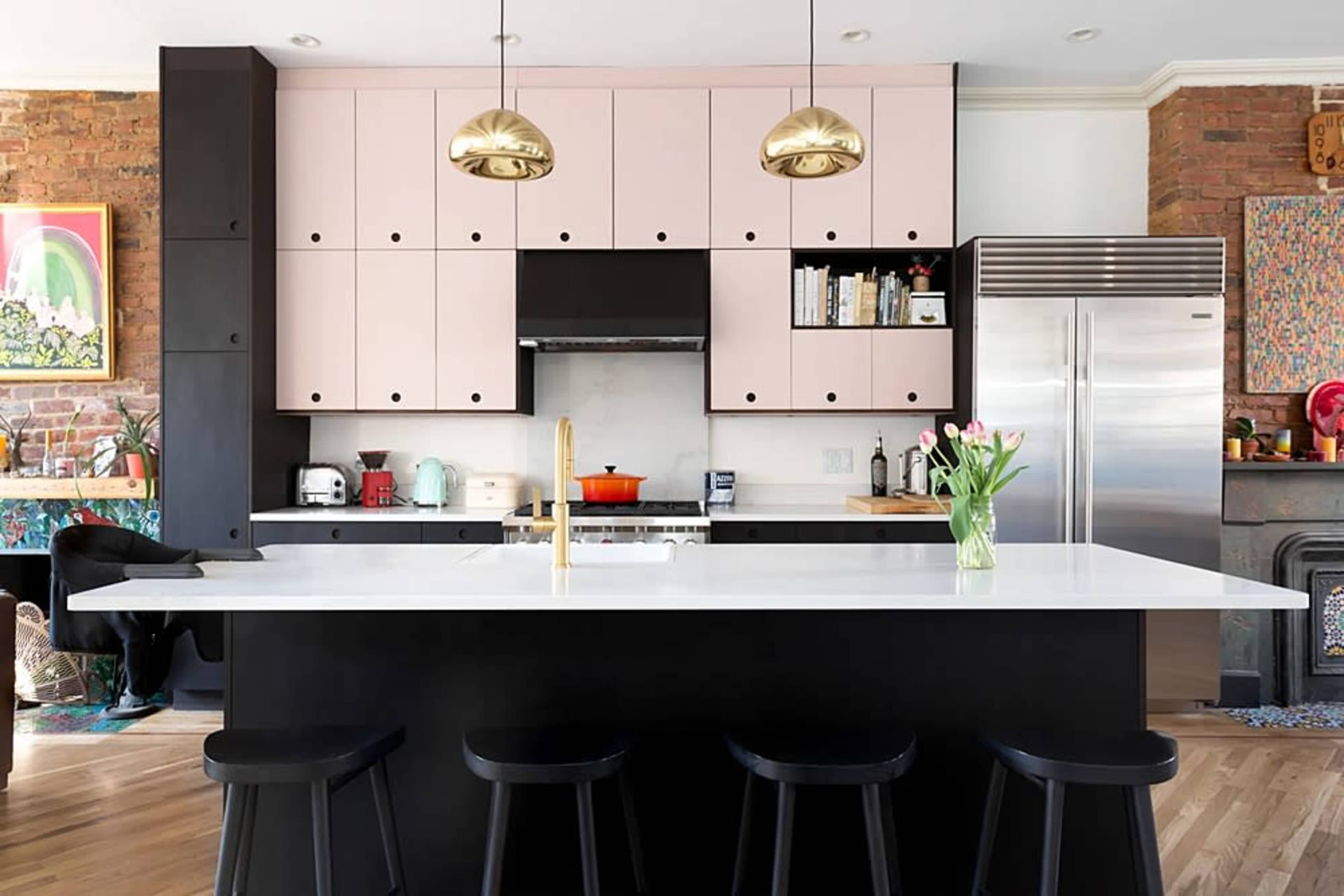 The image features a modern kitchen with pink and black cabinetry, a large white island with black stools, and gold pendant lights hanging above.