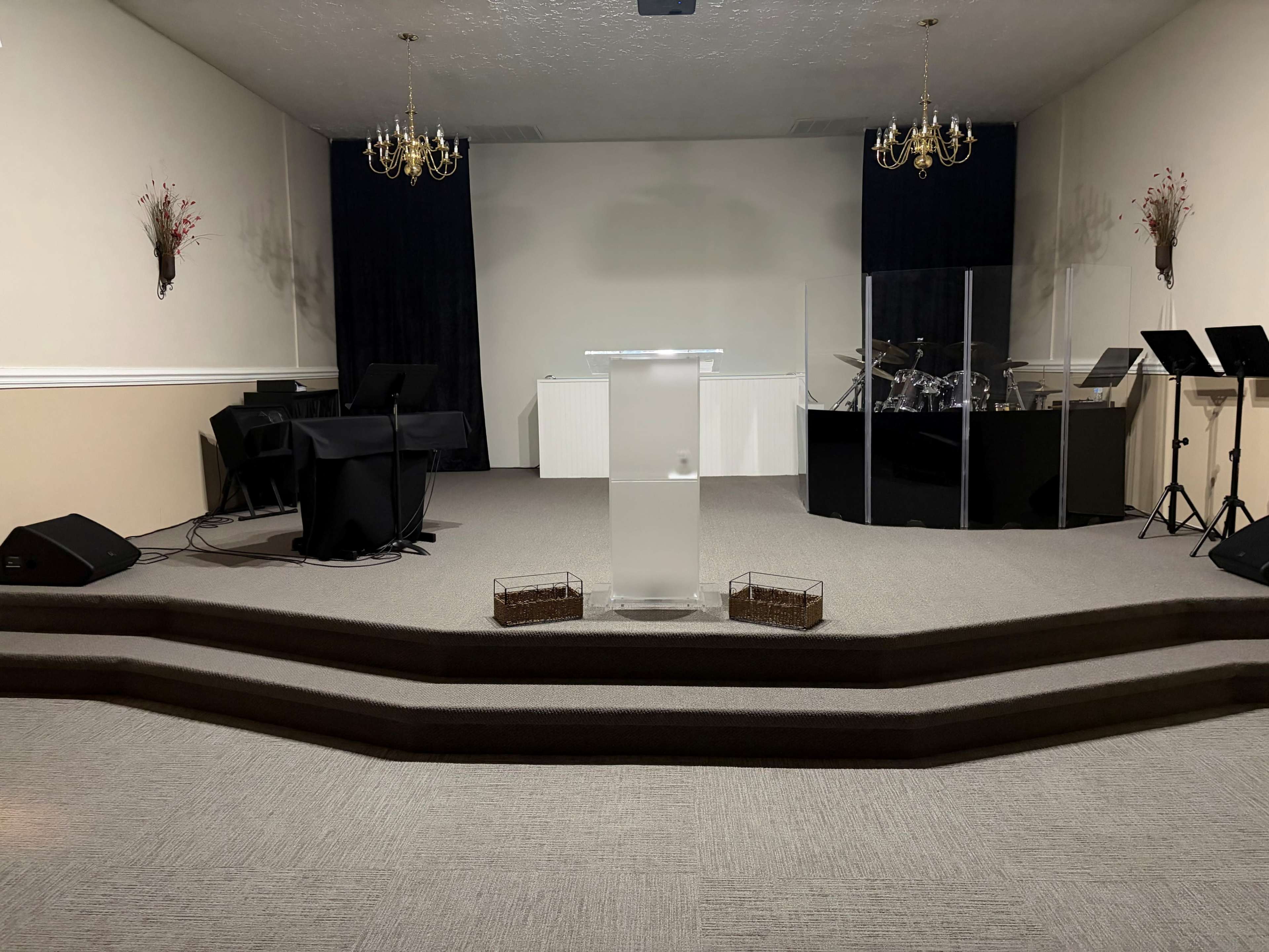 The image shows a stage area in a hall, featuring a transparent podium, musical instruments behind a glass enclosure, and several chairs arranged in front.