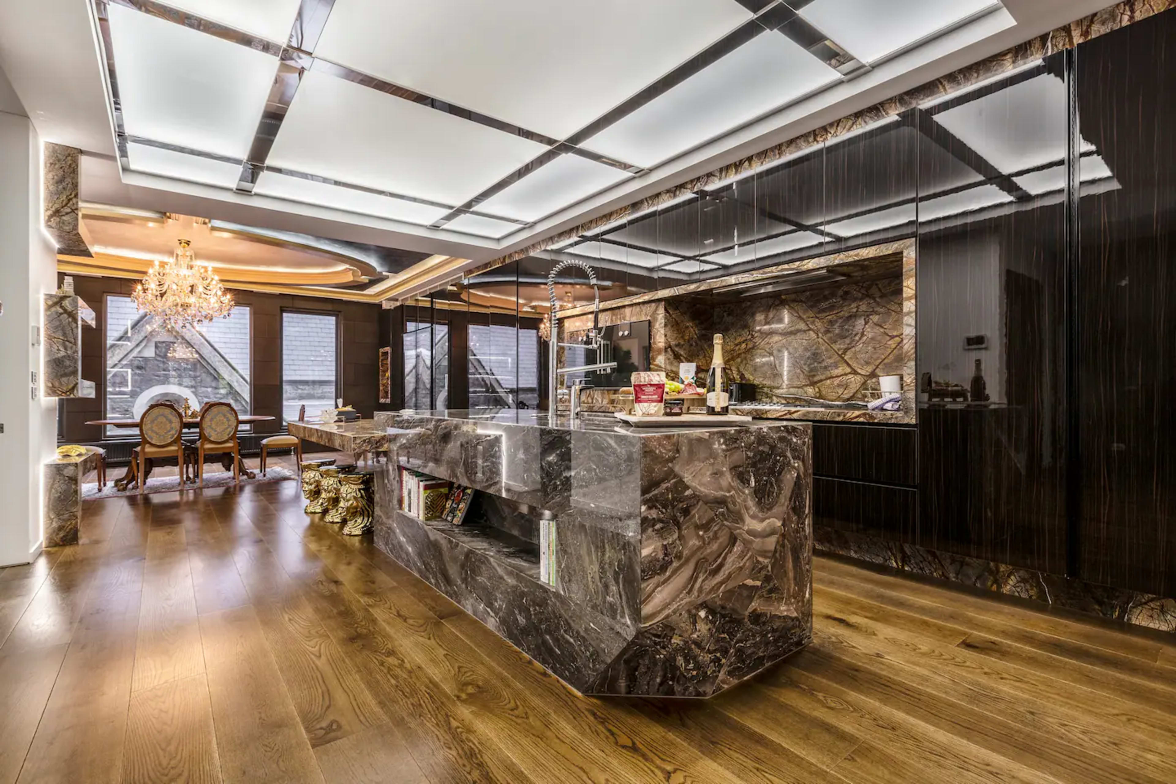 A modern kitchen with a large dark stone island and sleek black cabinetry, featuring a dining area with ornate chairs and a chandelier.