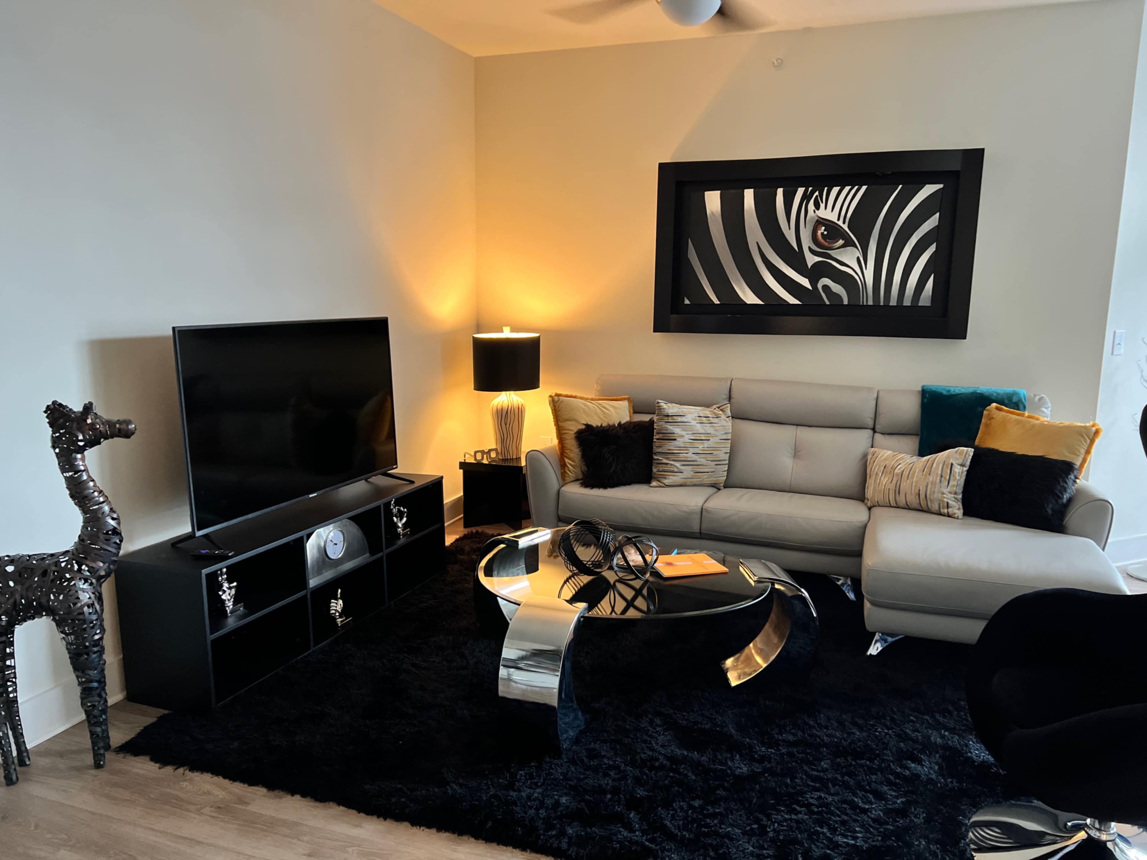 A modern living room featuring a light-colored sofa, a black entertainment unit with a large television, a glass coffee table, and decorative accents including a zebra-patterned artwork and a metallic sculpture.