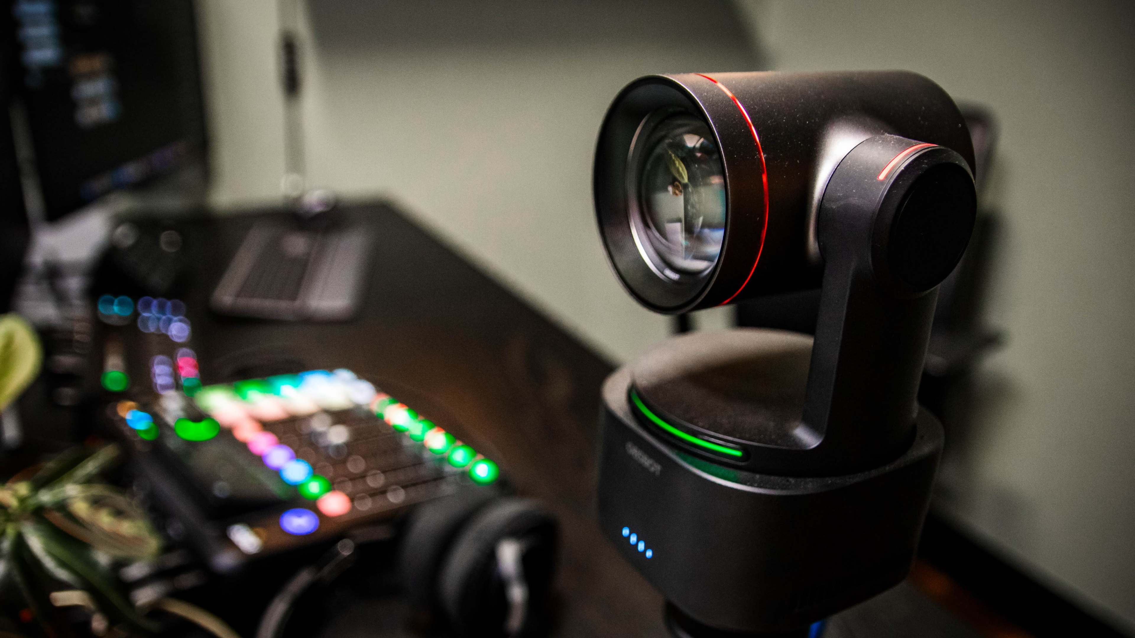 A stationary camera with a lens and red accents is positioned on a desk alongside a colorful control panel and a computer keyboard.
