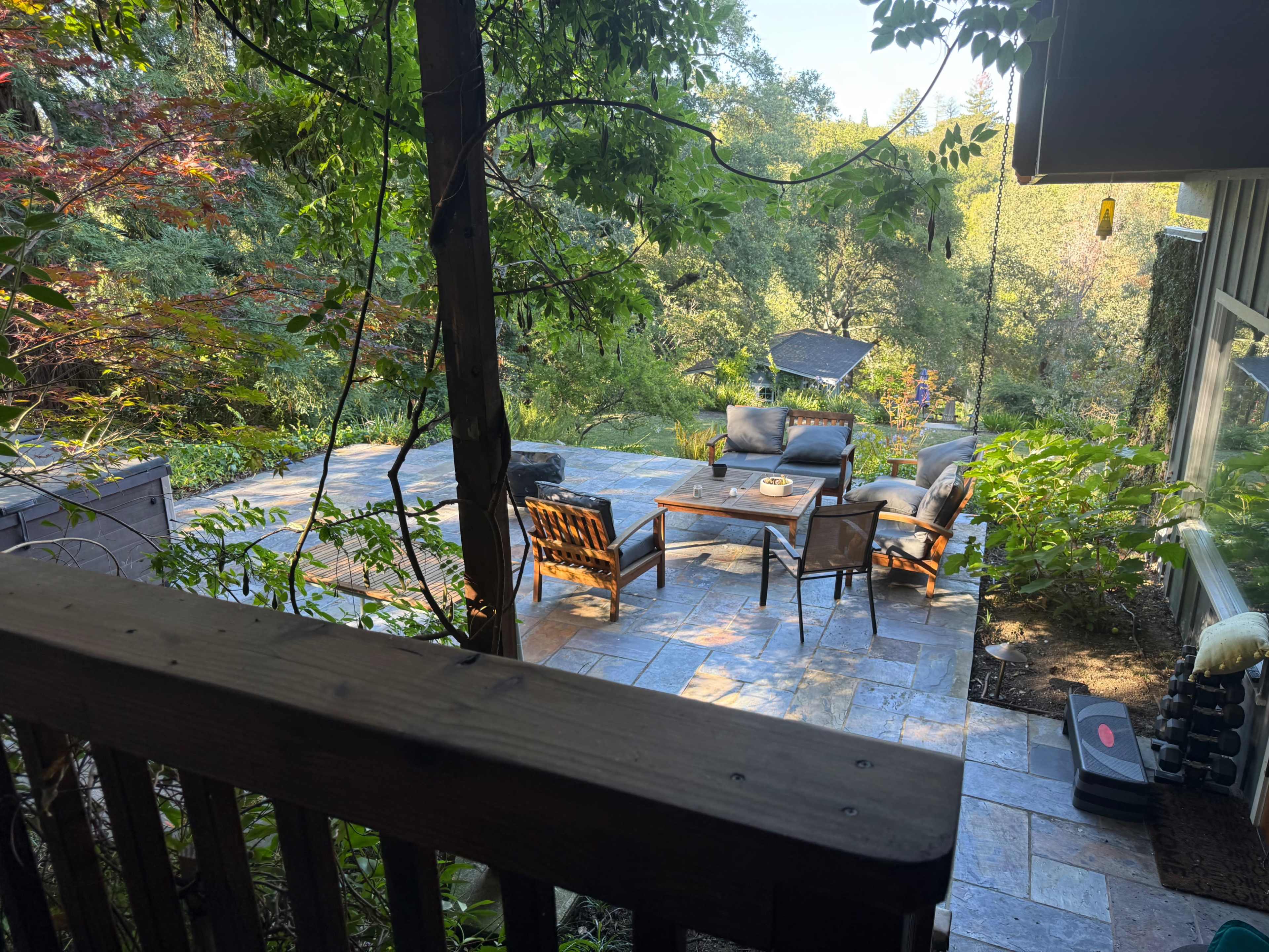 The image shows a stone patio area surrounded by greenery, featuring a set of outdoor furniture, including chairs and a table.