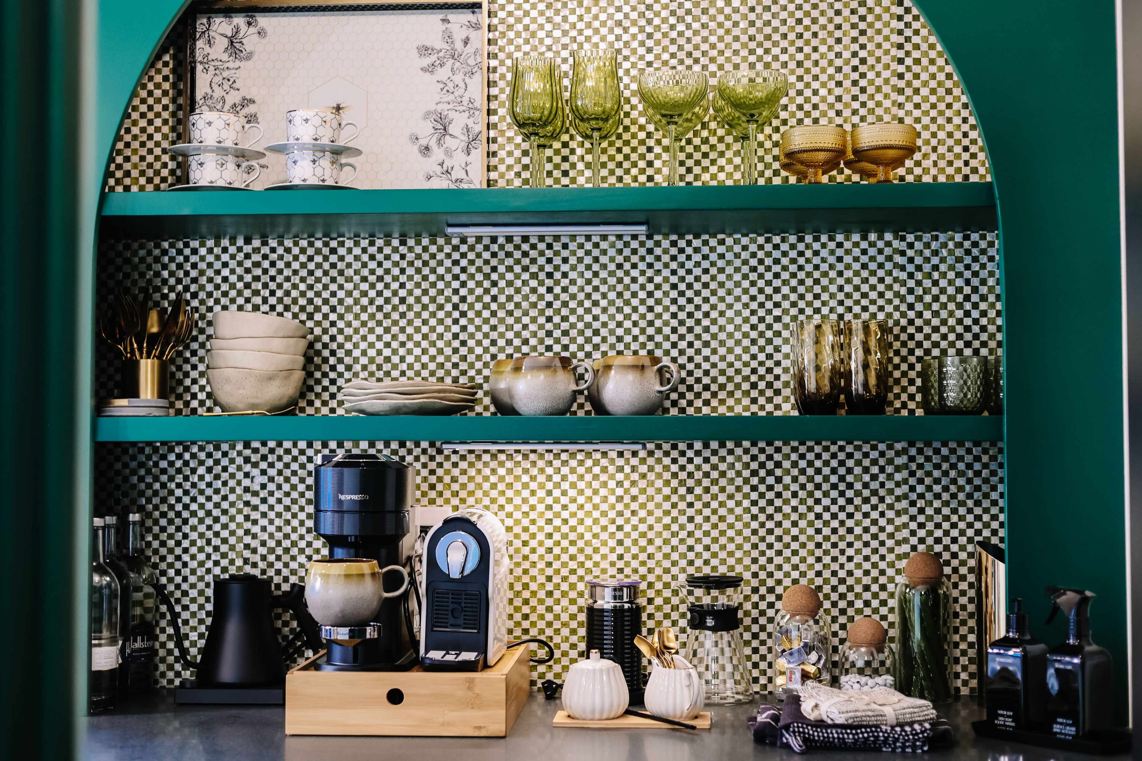 A kitchen shelf displays various dishes, glassware, and a coffee machine, set against a patterned backsplash.