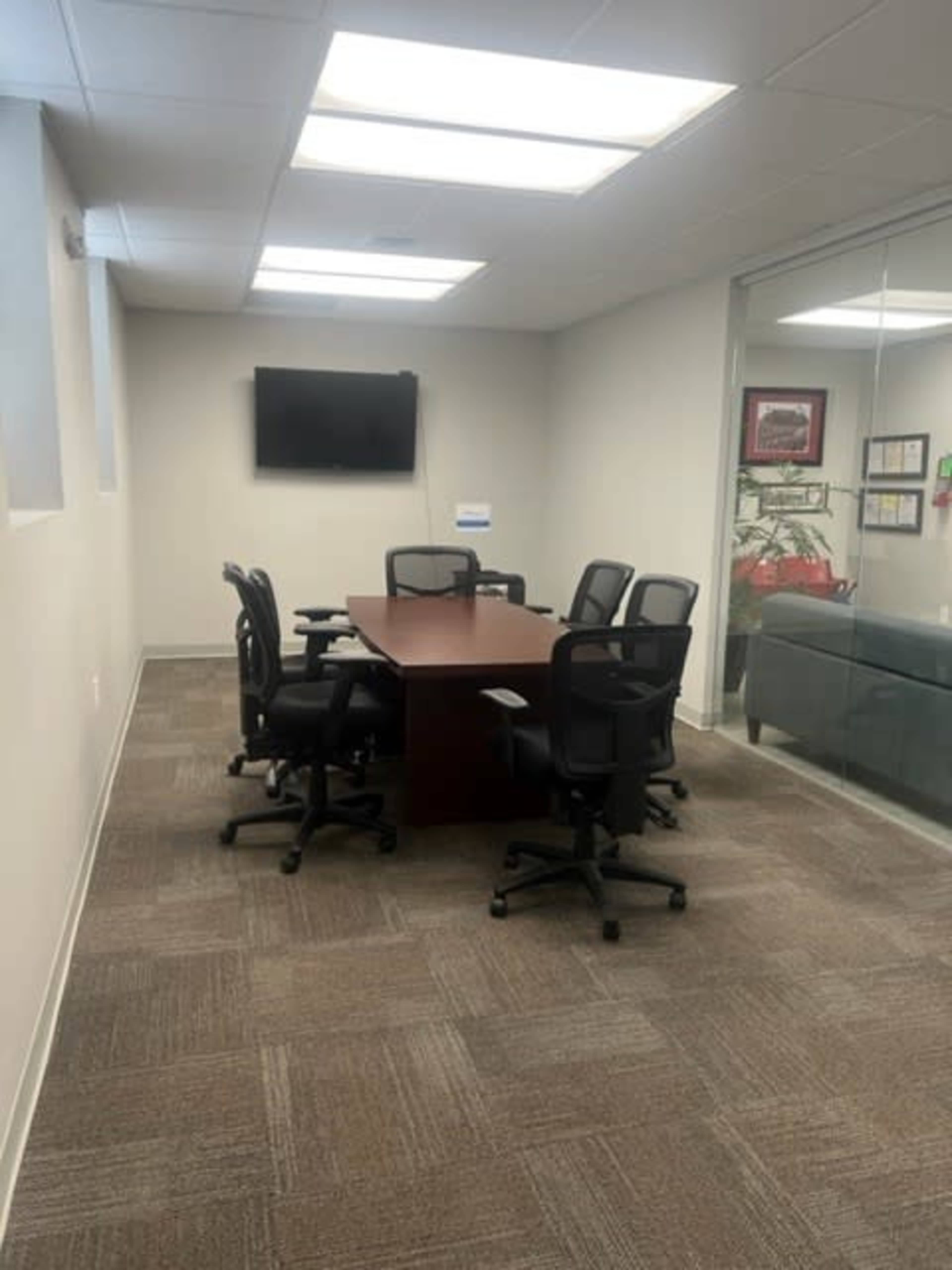 The image shows a conference room with a rectangular table surrounded by chairs, a wall-mounted television, and a glass wall.