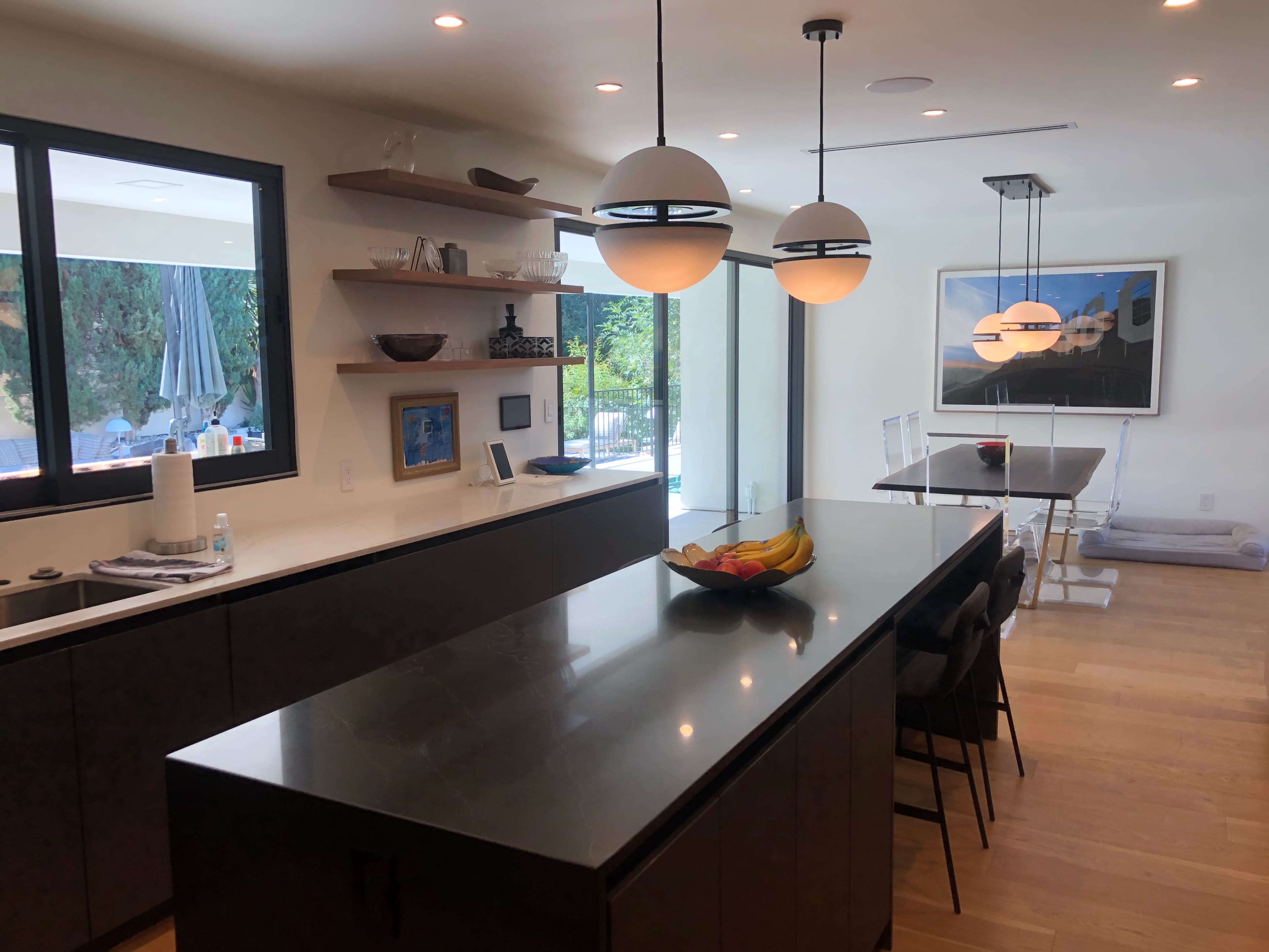 A modern kitchen features a dark island with a bowl of fruit, shelves displaying decor, and large windows overlooking a patio with a pool.