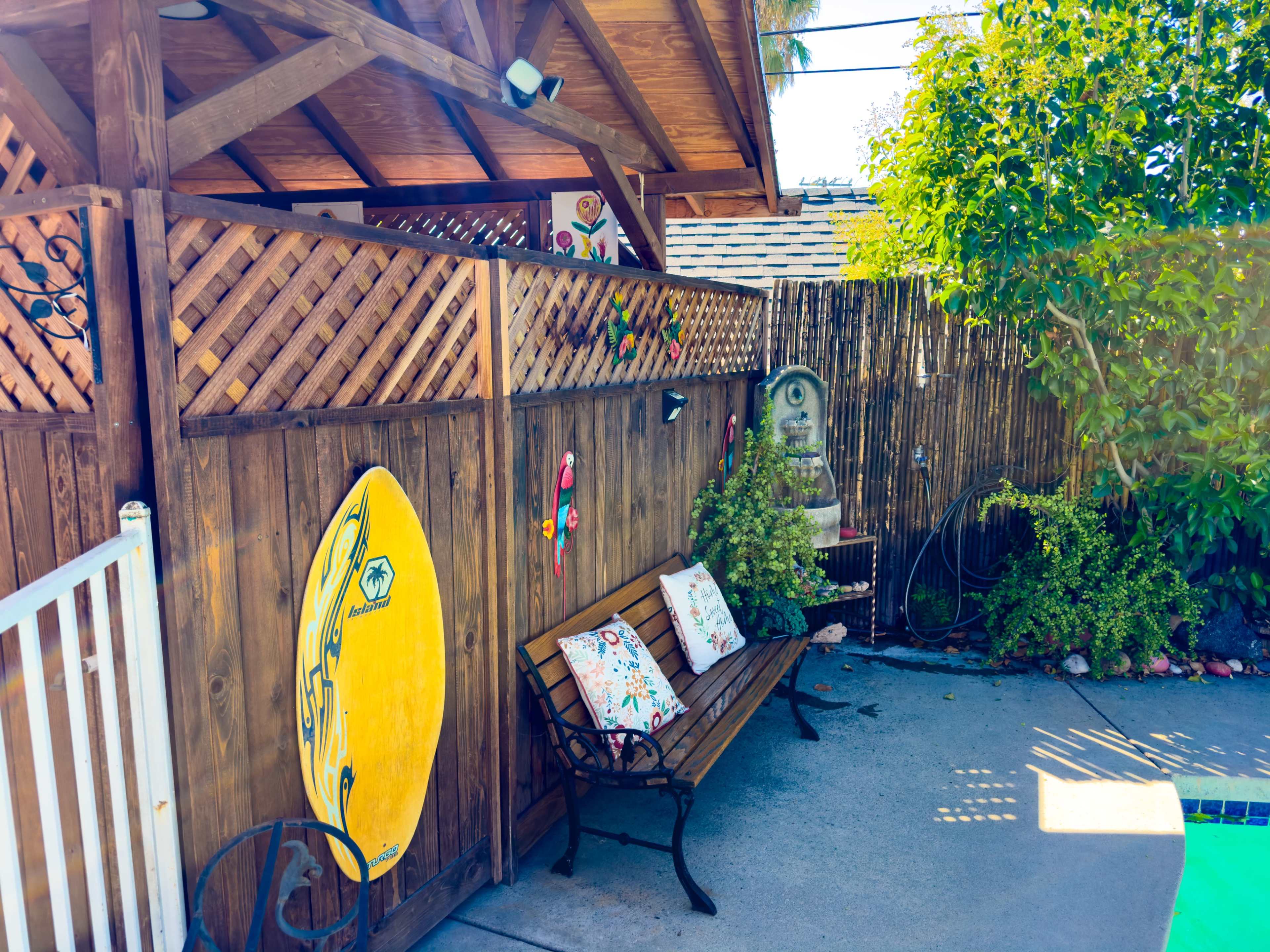 The scene features a wooden bench with colorful pillows positioned beside a surfboard mounted on a fence, near a pool surrounded by greenery.