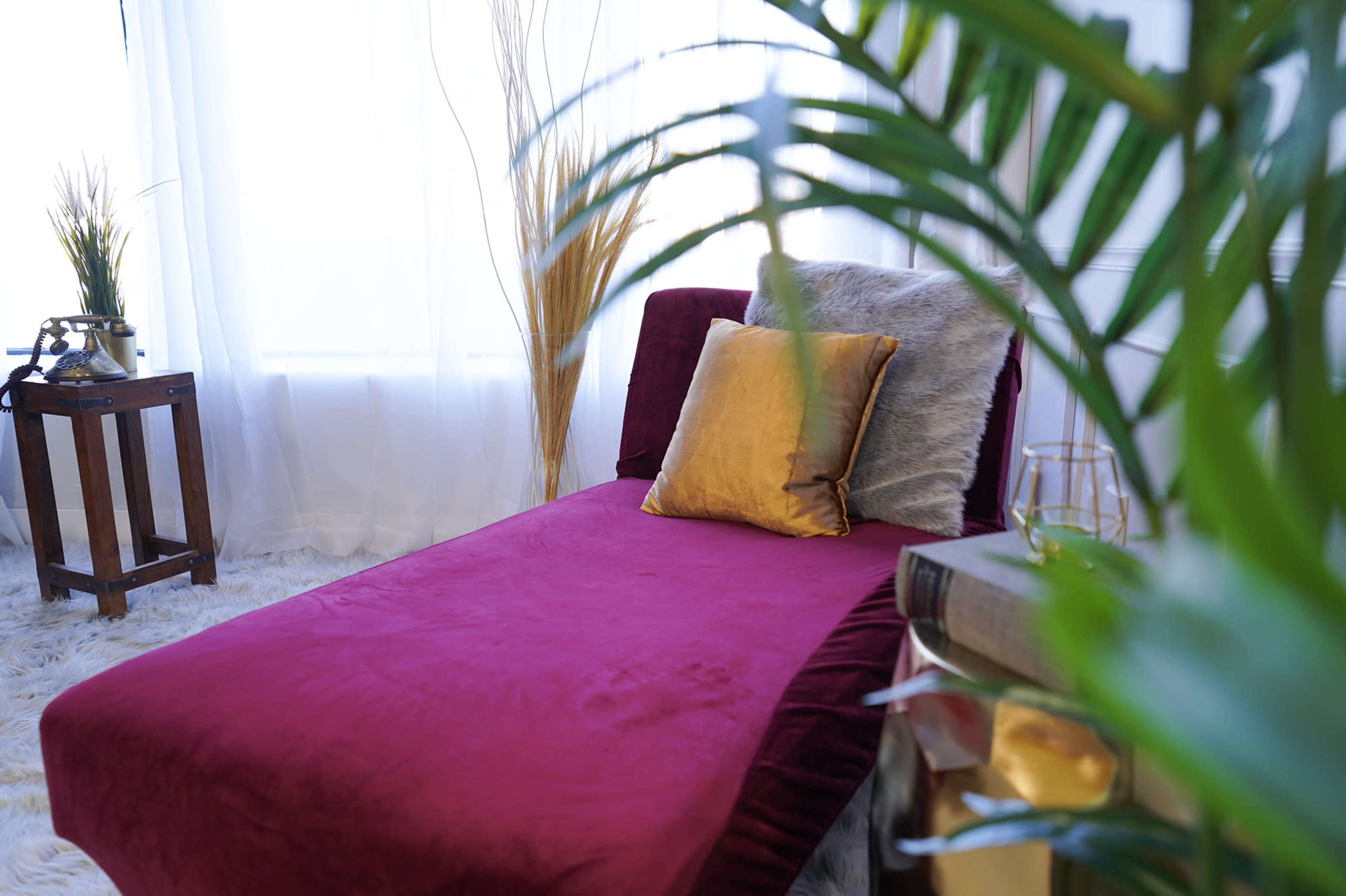 A burgundy daybed adorned with a gold pillow is positioned near a window, surrounded by indoor plants and decorative greenery.