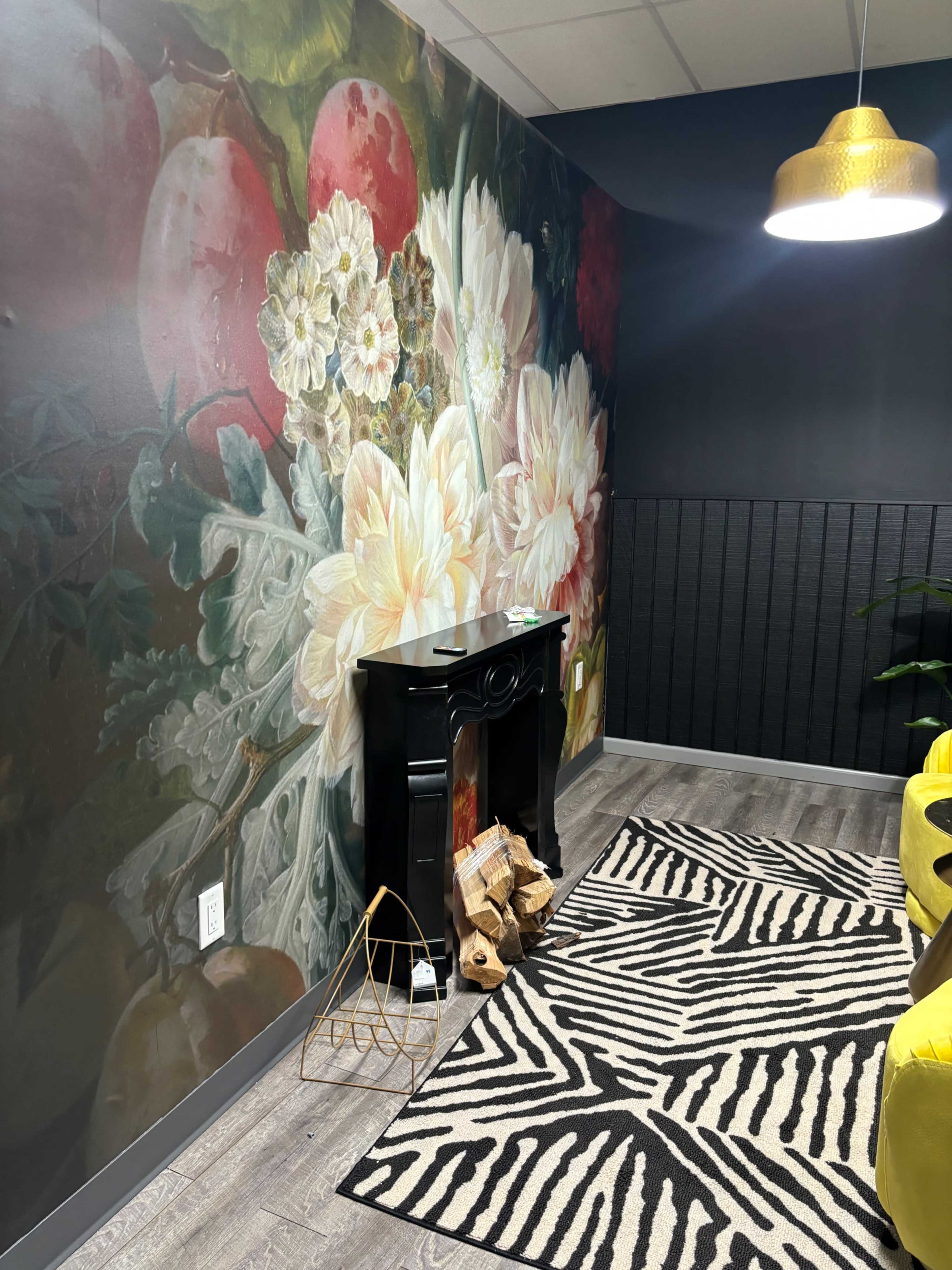 A corner of a room featuring a large floral mural, a black faux fireplace with stacked logs, and a yellow sofa, all set against a black wall with a patterned rug.