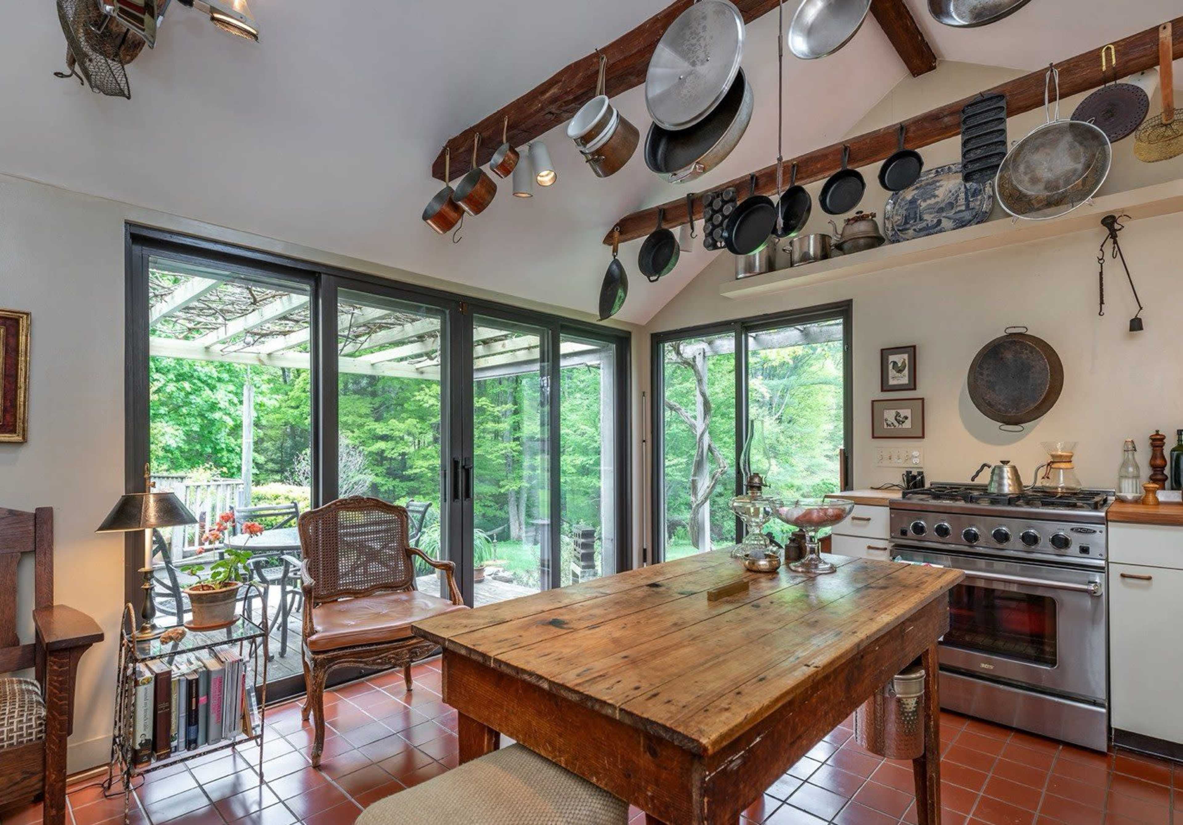 The image shows a kitchen with a wooden table, several cooking pots hanging from the ceiling, and large windows overlooking a green outdoor area.