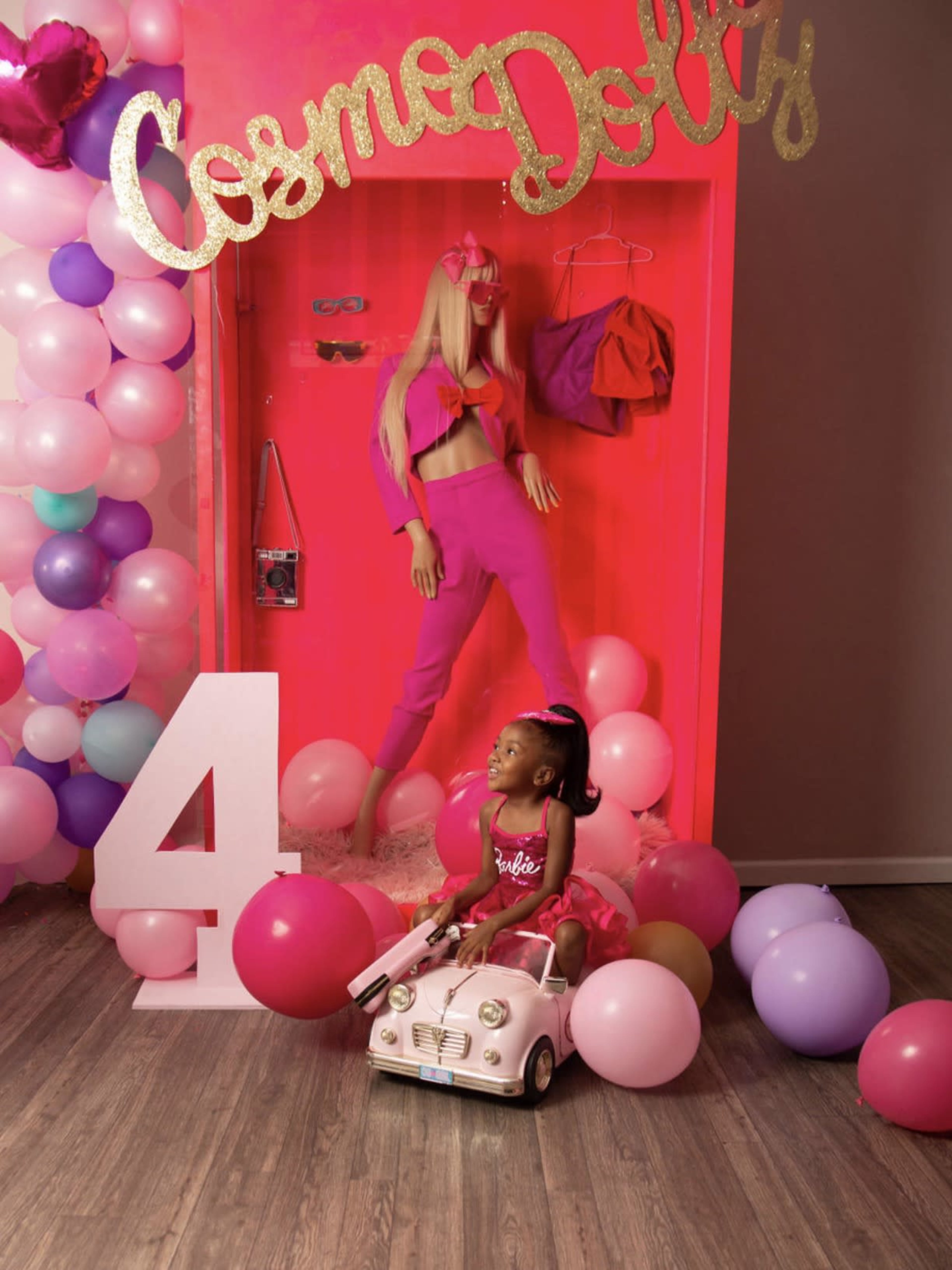 A young girl in a pink dress sits in a toy car surrounded by colorful balloons and a mannequin dressed in pink, with the words "Cosmo Dolly" displayed above.