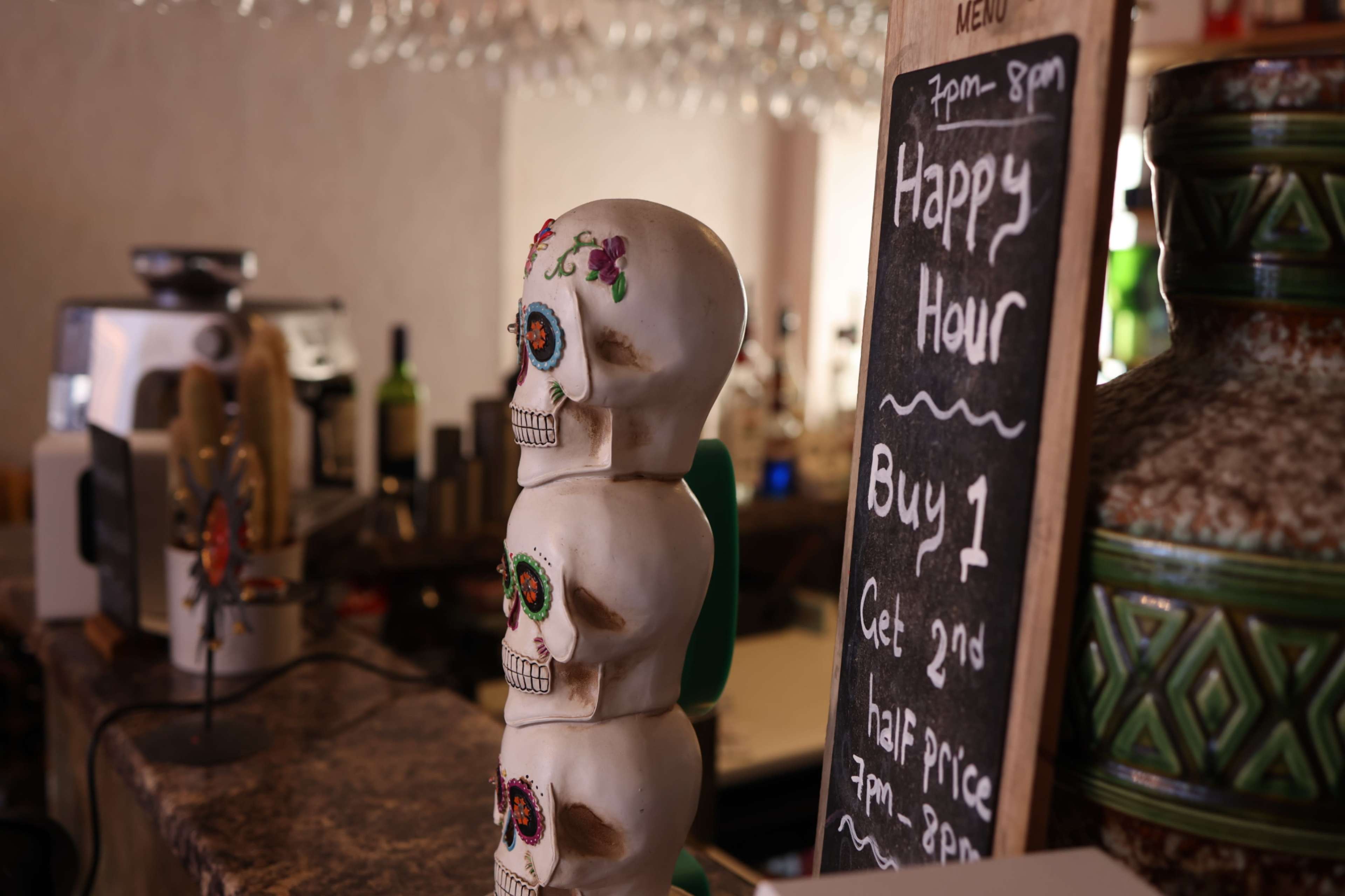 Three decorative skulls are displayed next to a chalkboard sign advertising a happy hour promotion at a bar.
