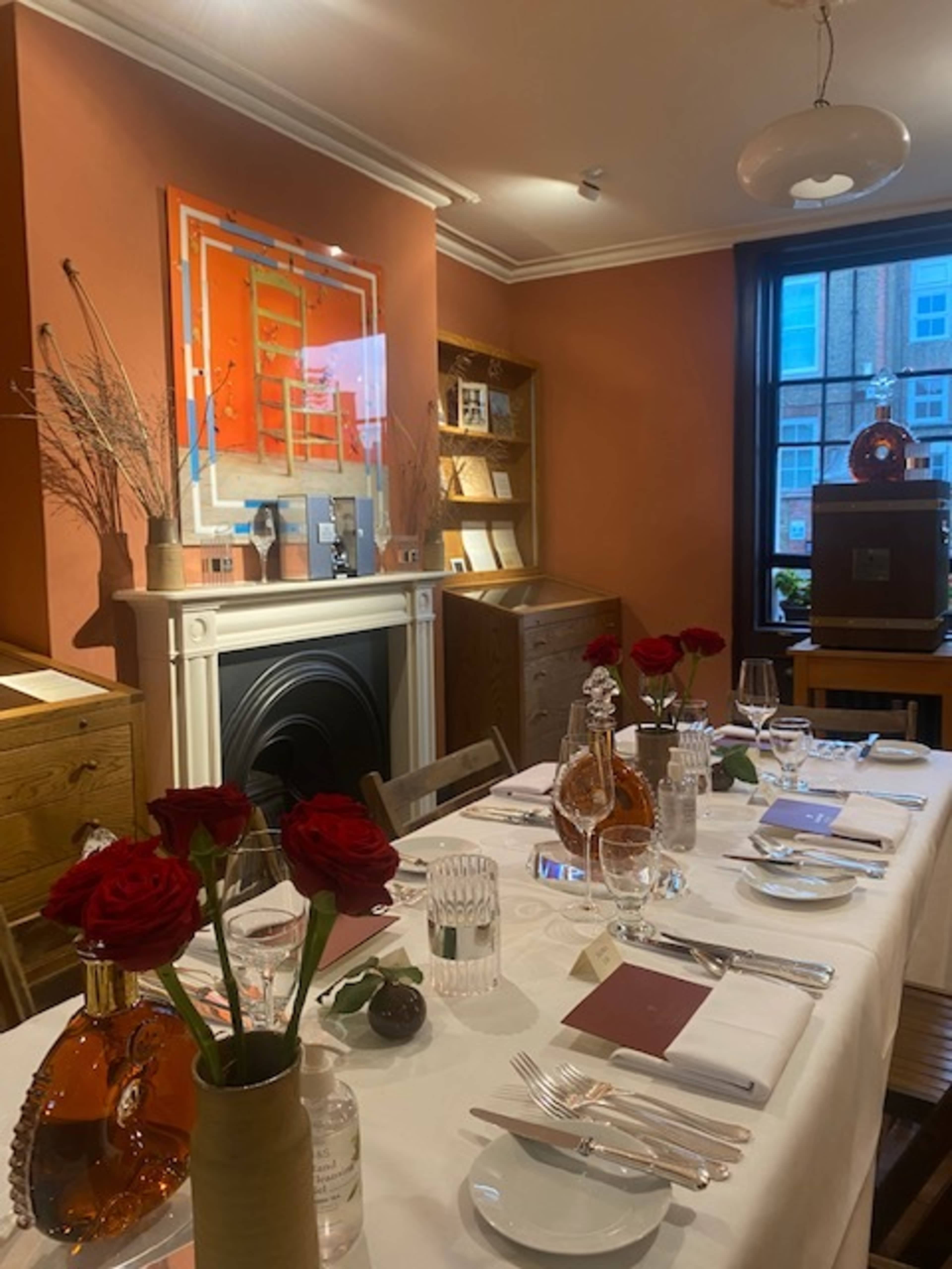 The image shows a elegantly set dining table with red roses, fine glassware, and decorative items in a stylish room featuring orange walls and a fireplace.