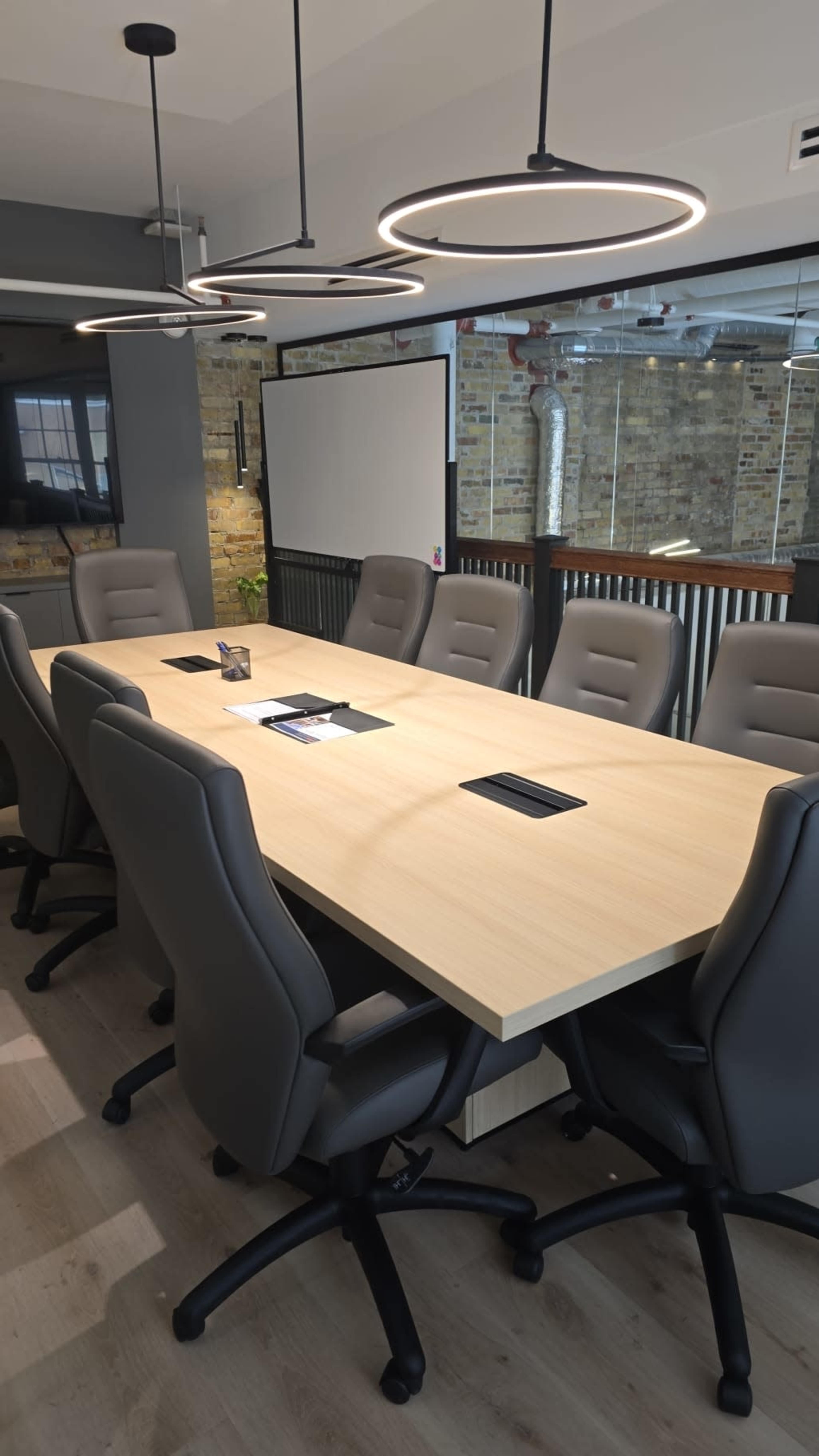 A conference room features a long wooden table surrounded by ergonomic chairs, with modern lighting fixtures hanging overhead.