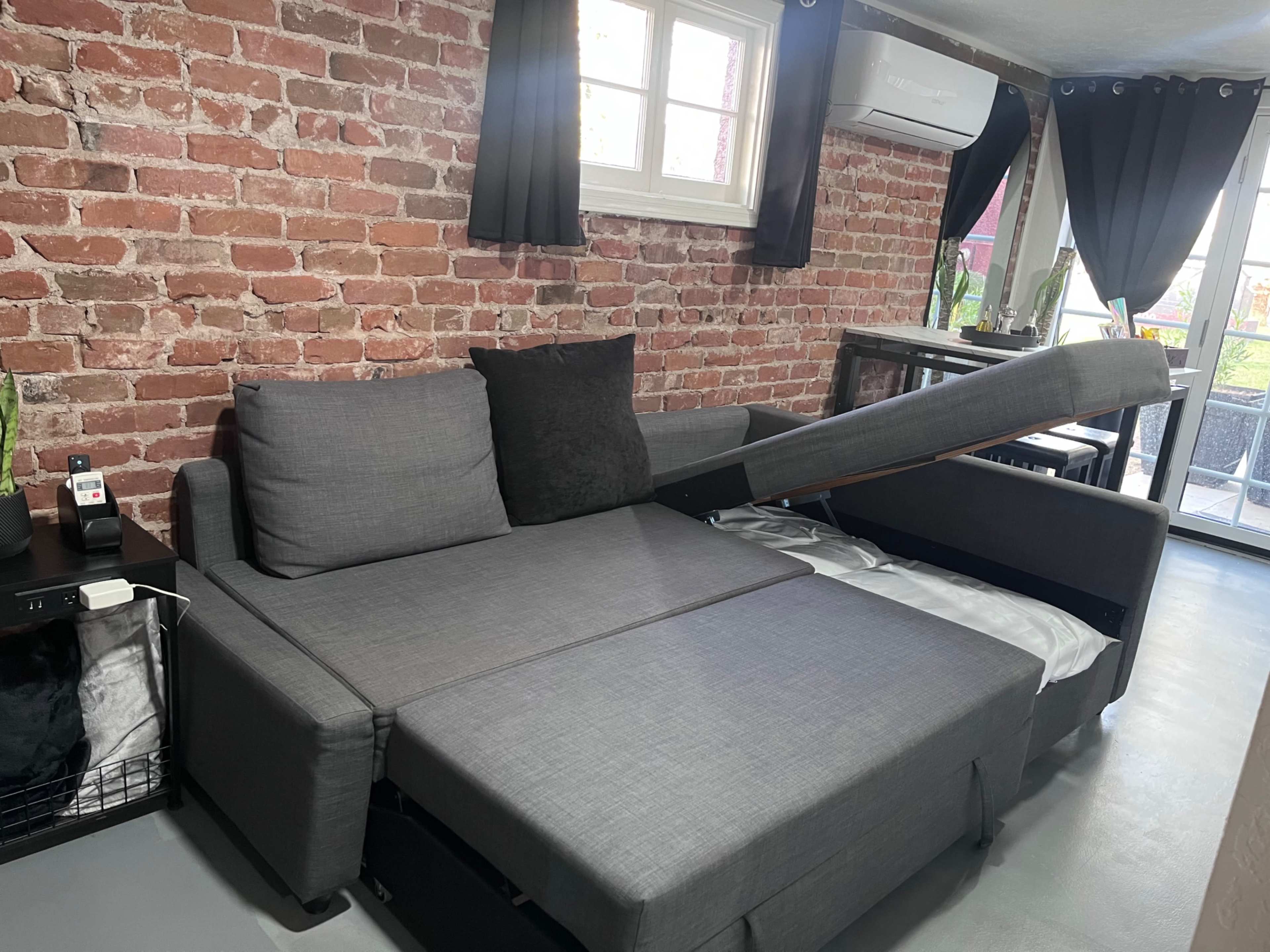 The image shows a gray sectional sofa with a pull-out bed mechanism against a brick wall, accompanied by black curtains and a small table in the background.