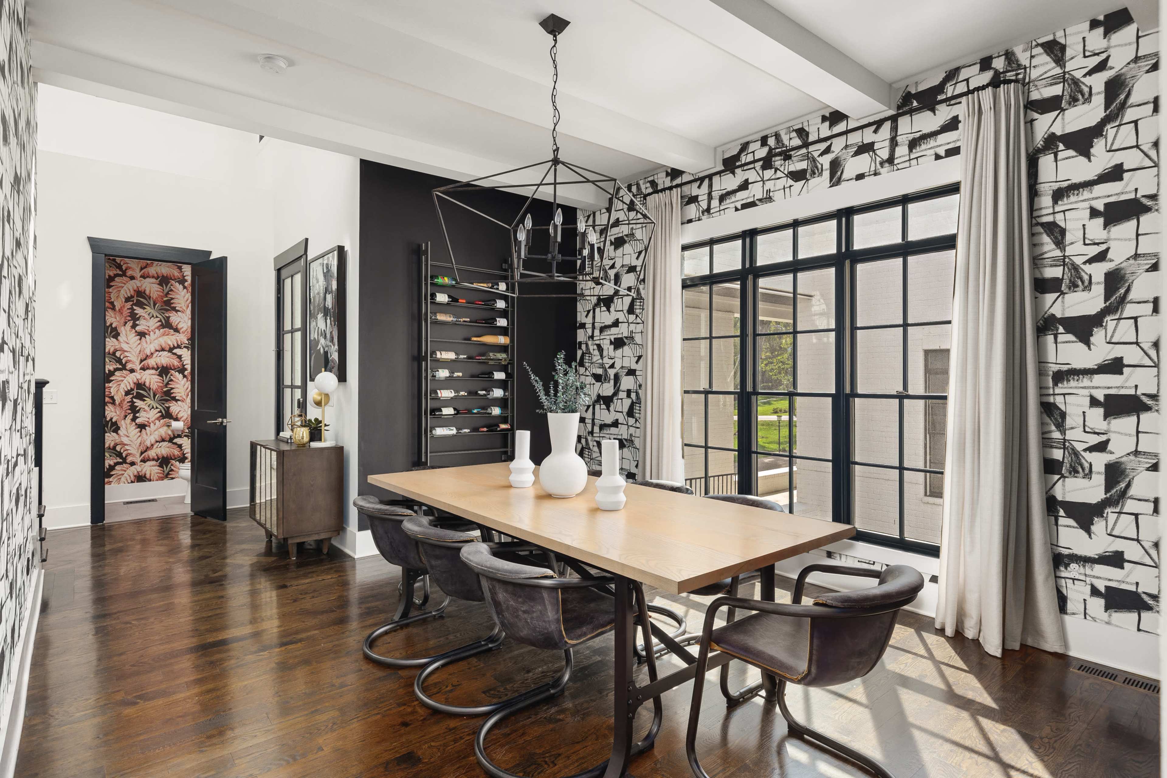 The image shows a modern dining area with a large wooden table, surrounded by black chairs, and features patterned wallpaper and large windows allowing natural light.