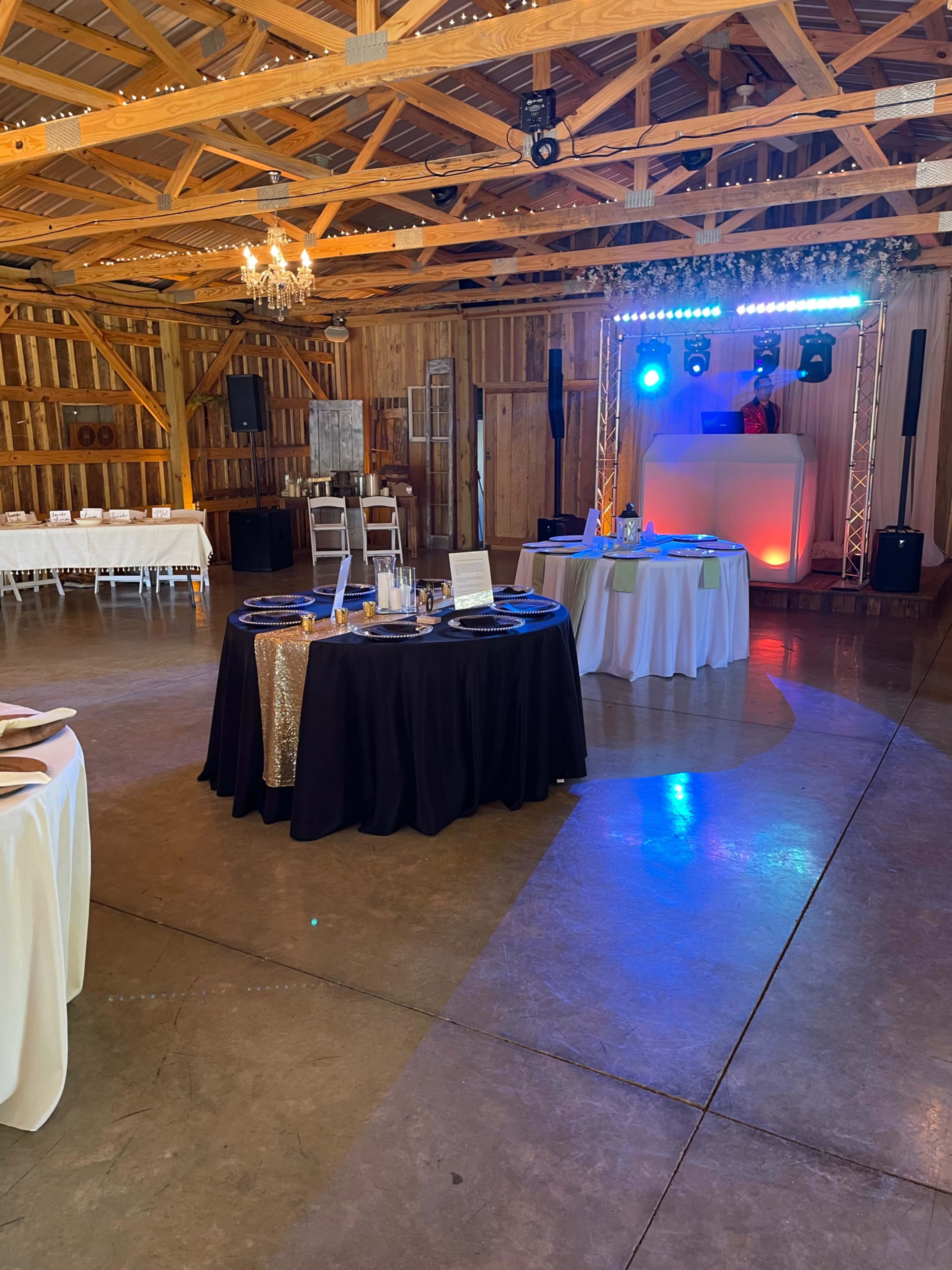 The interior of a barn venue features tables draped in white and black linens, with lighting and a performance area at one end.