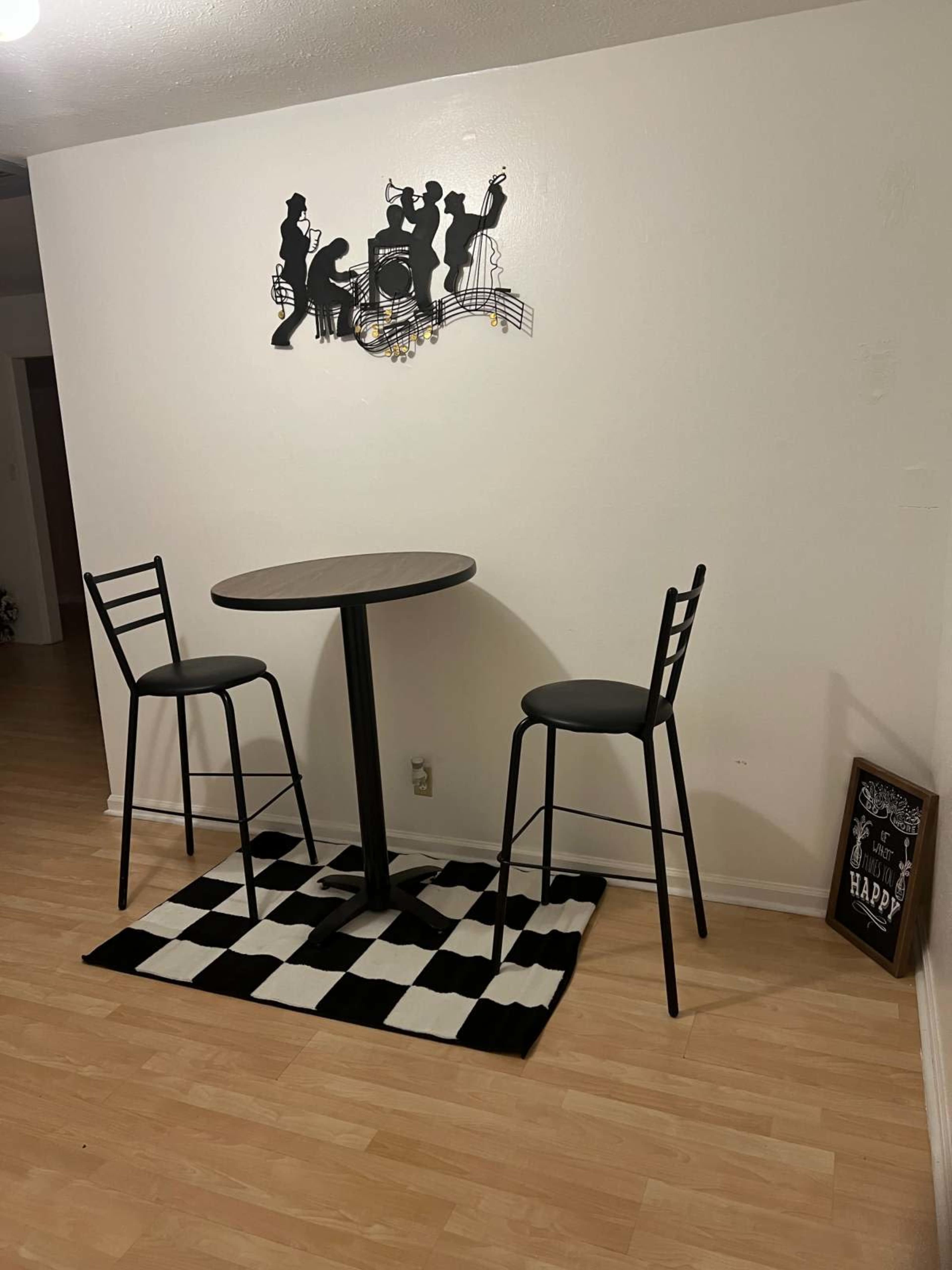 A small dining area features a round table with two high-backed stools on a black and white checkered rug, accompanied by a musical-themed wall decoration and a framed sign.