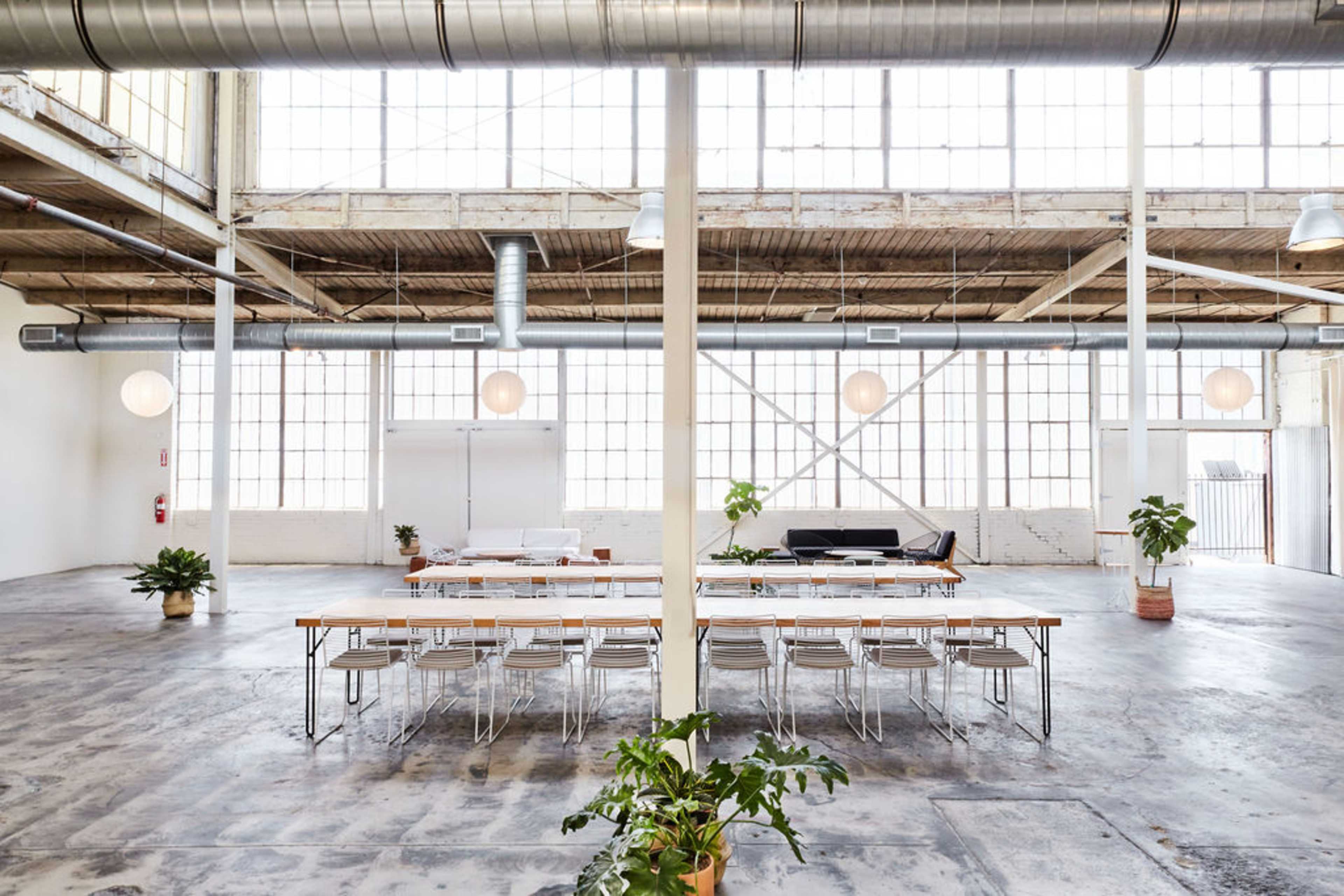 The image shows a spacious, industrial-style room with large windows, featuring a long dining table surrounded by chairs, and plants in decorative pots.