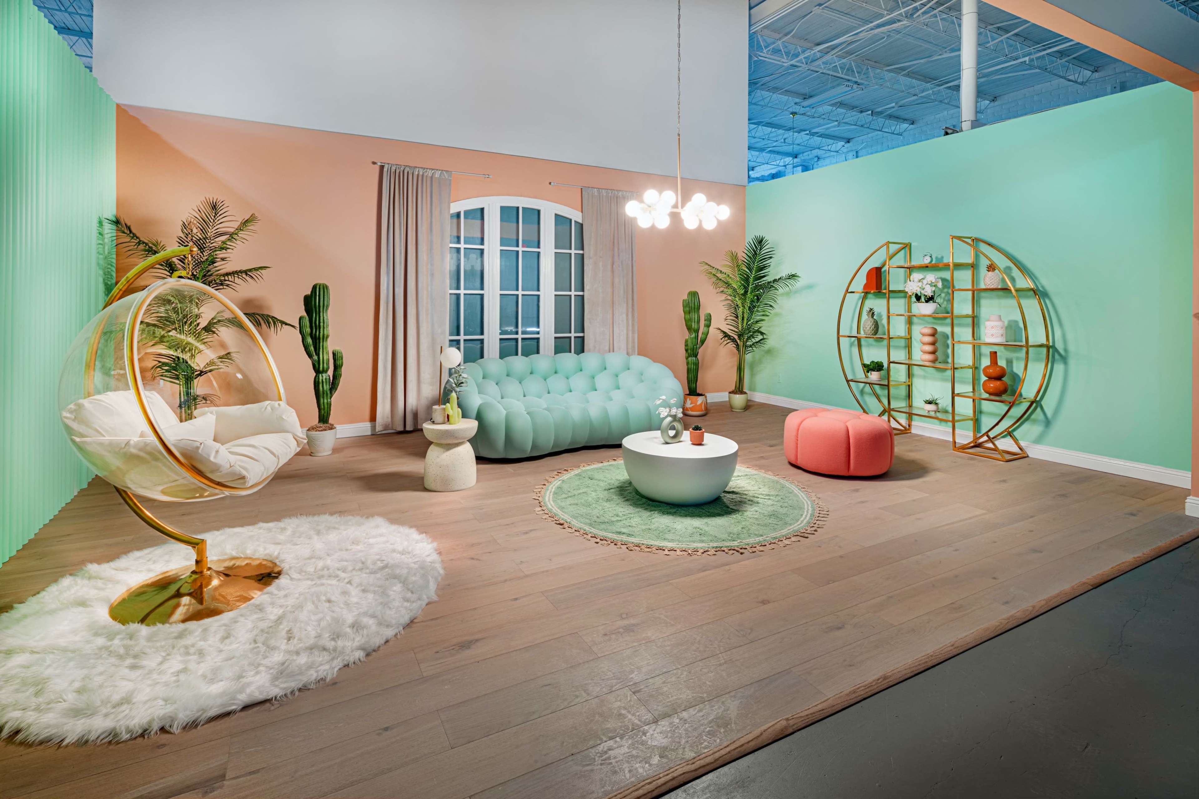 A brightly colored room features a modern lounge area with a bubble-shaped chair, a tufted turquoise sofa, a round coffee table, and decorative accents on a gold shelving unit.