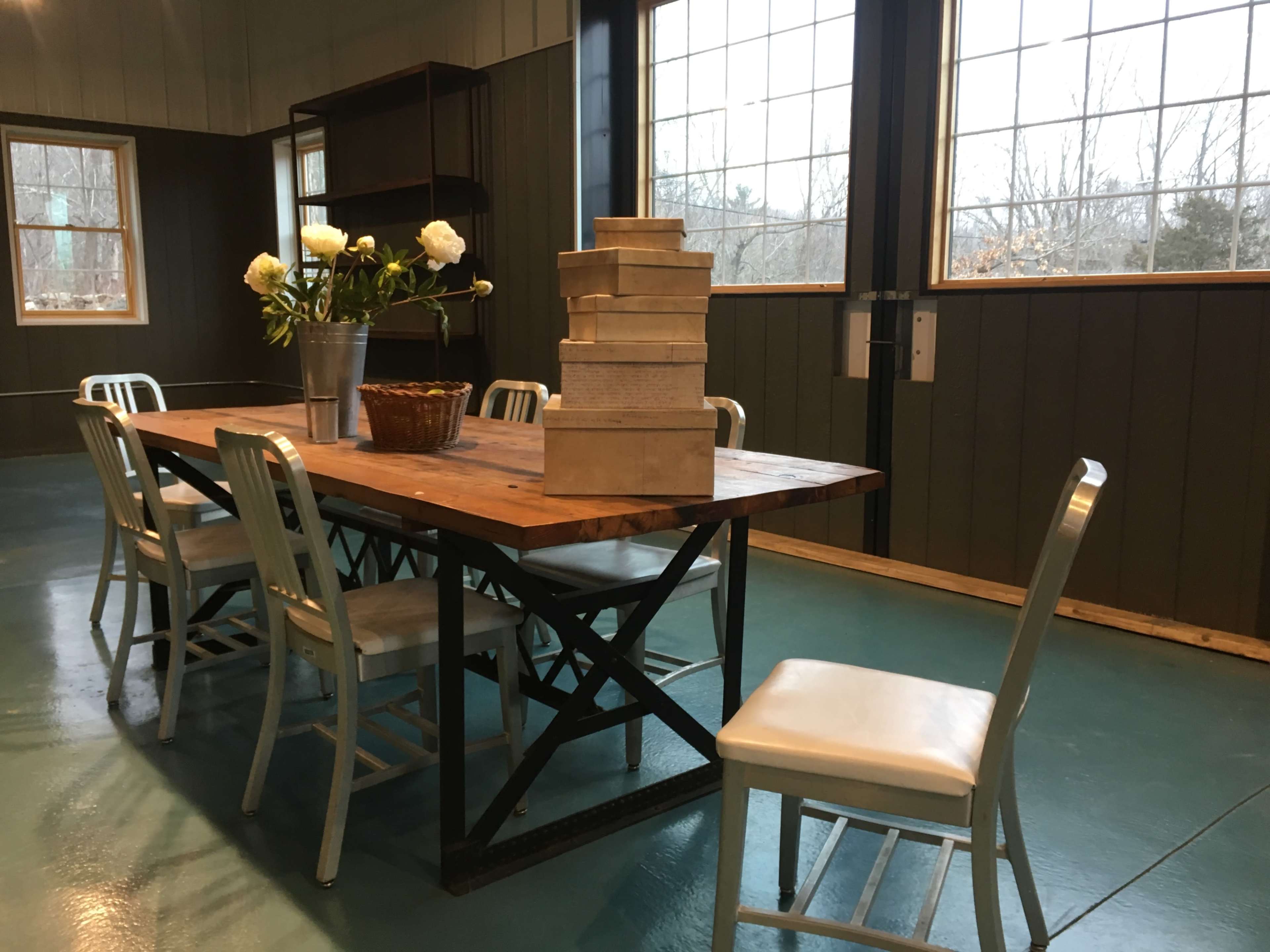 A wooden table with a stack of boxes and a vase of flowers is surrounded by six metal chairs in a room with large windows.