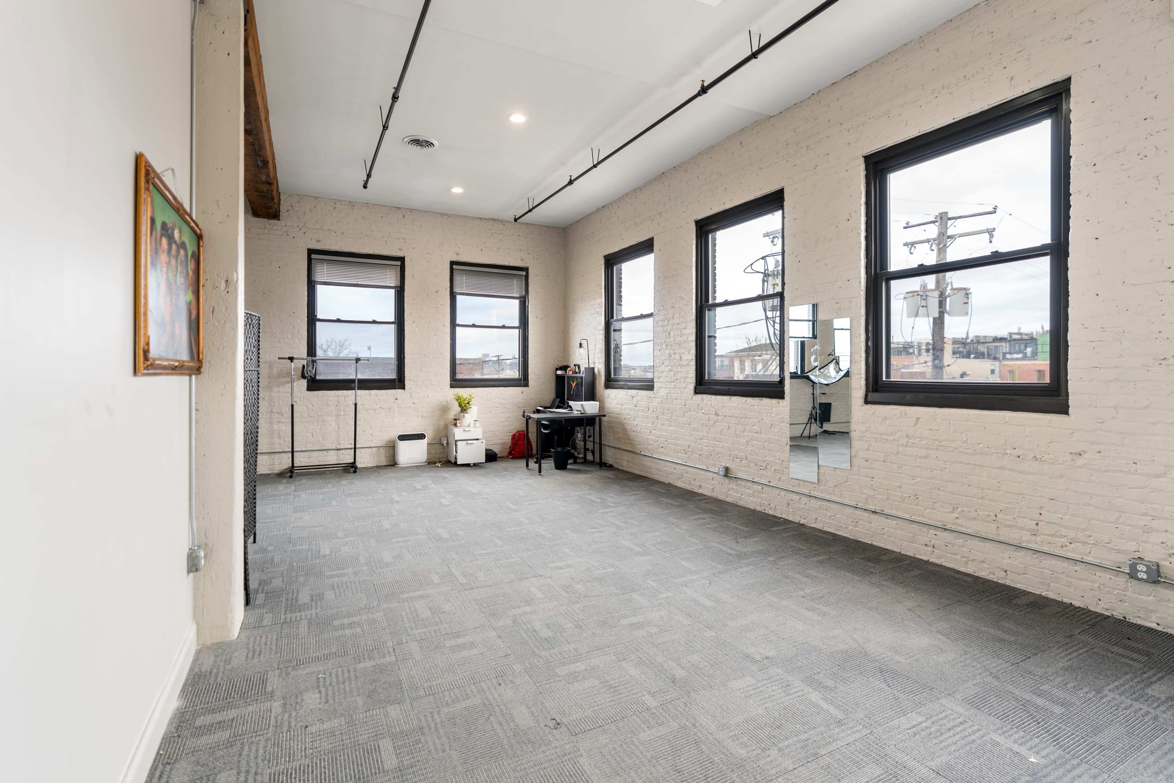 A spacious room with four windows, light-colored brick walls, and gray carpet, featuring an area with a desk and a mirror.