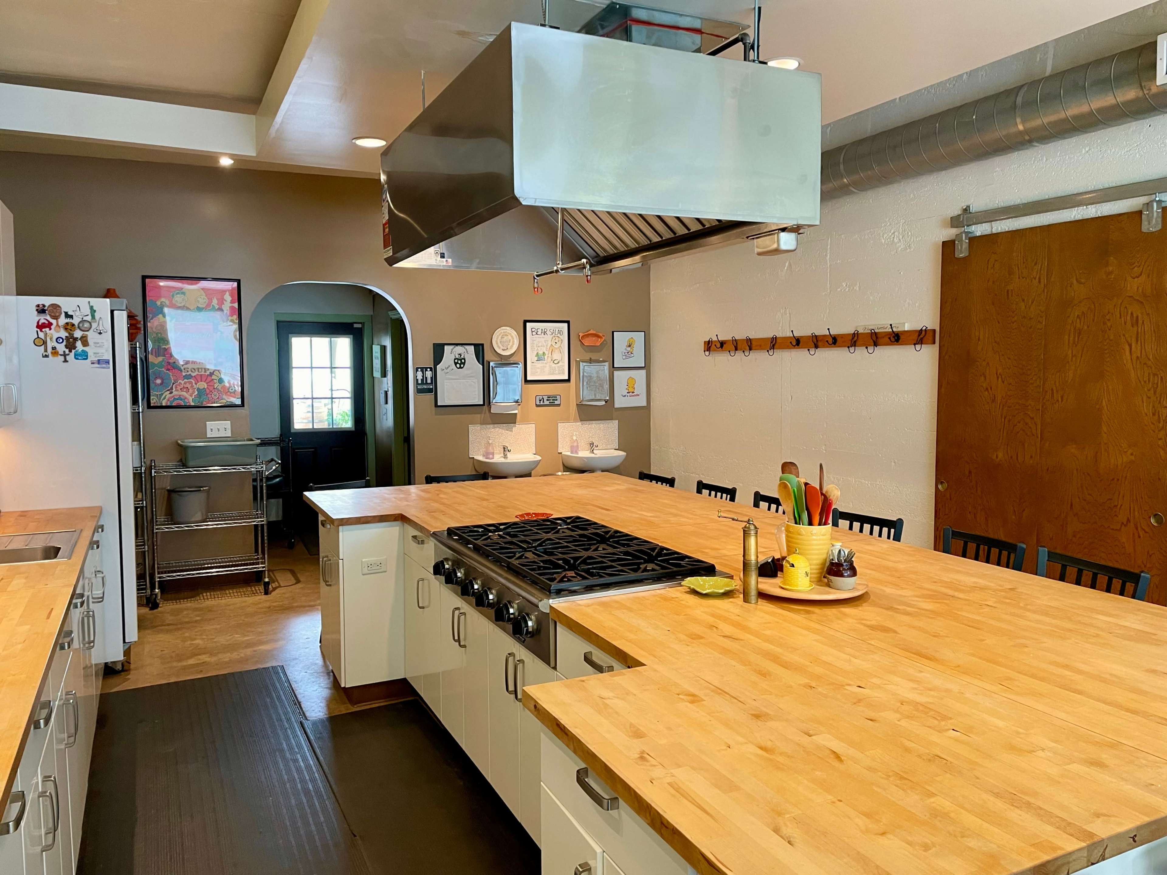 A modern kitchen features a central island with a gas stove, wooden countertops, and white cabinetry, with an archway leading to a doorway in the background.