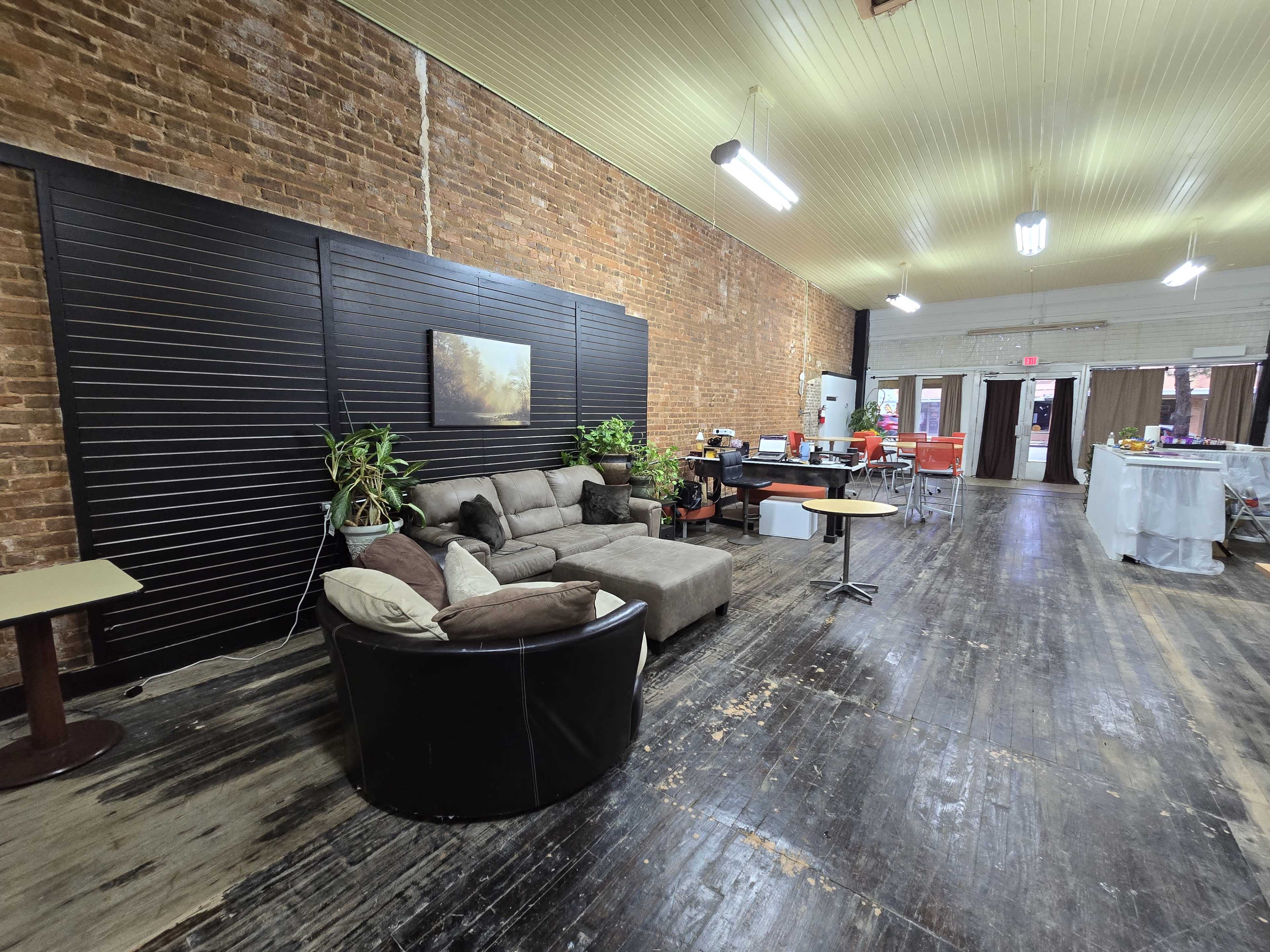 The image shows a spacious, renovated room with exposed brick walls, a seating area featuring a dark sofa and a circular chair, and a kitchen area in the background.