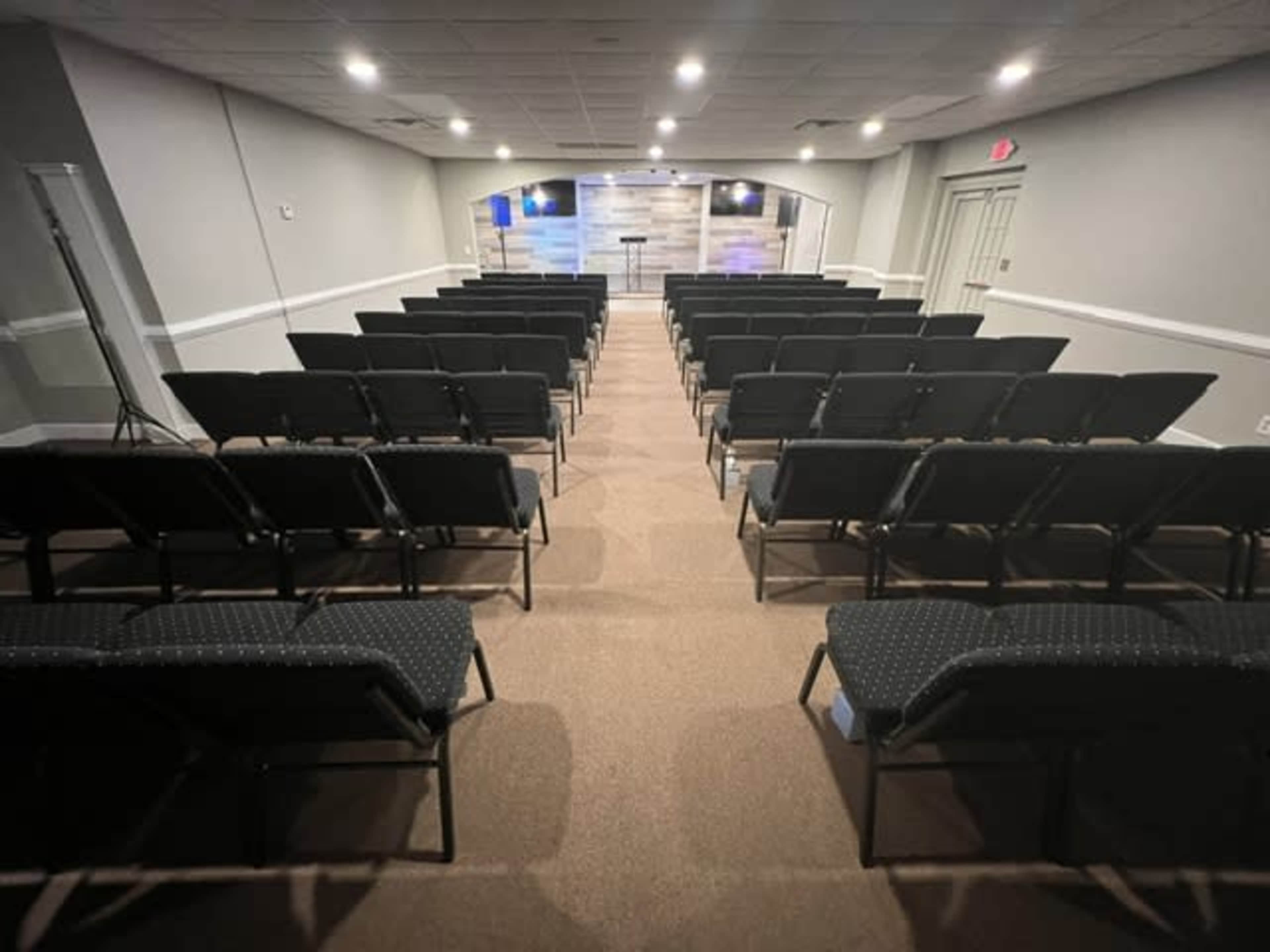 An empty auditorium with rows of black chairs facing a stage at the front, equipped with a podium and screens.