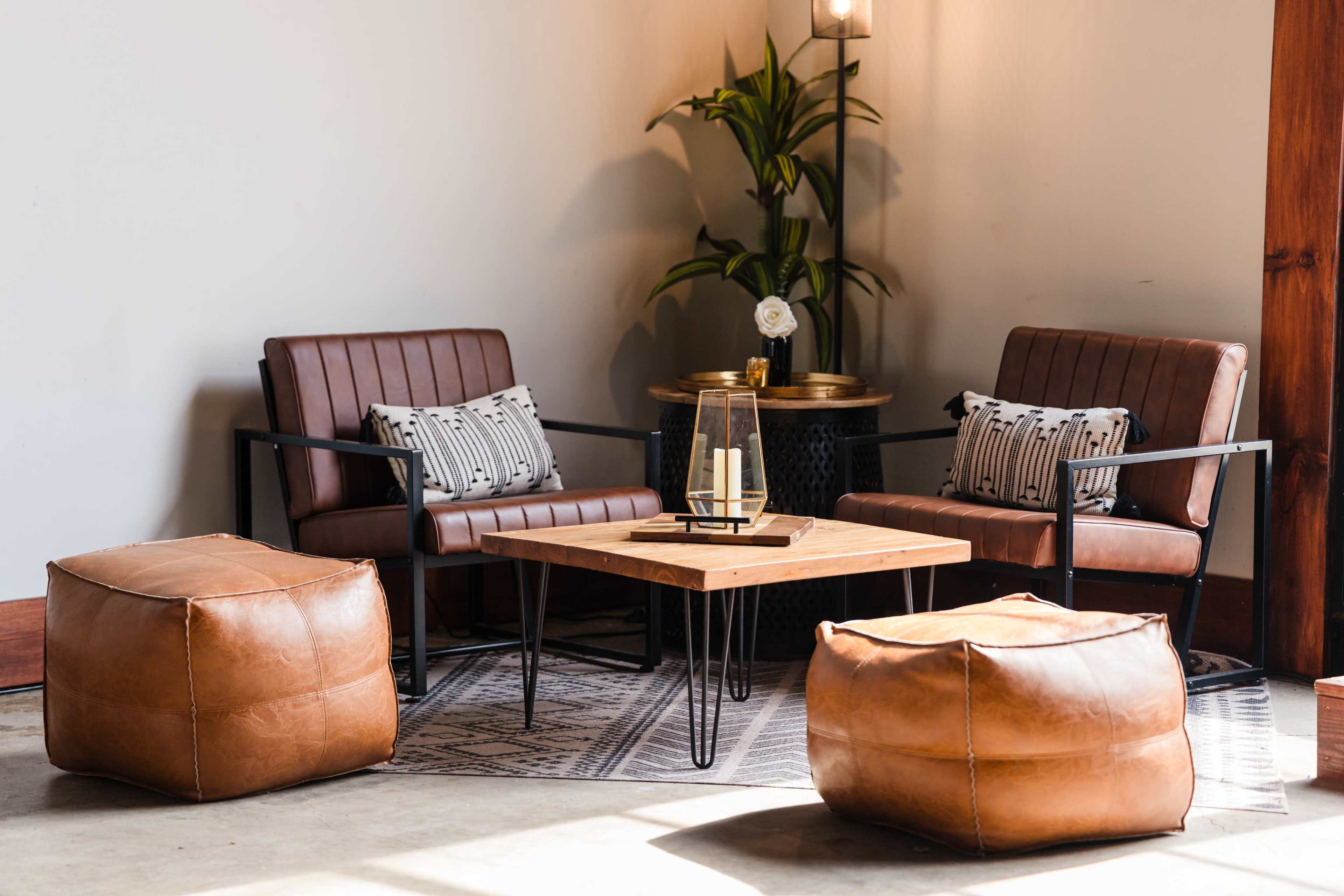 The image features a modern lounge area with two brown upholstered chairs, a wooden coffee table, and two leather ottomans arranged on a patterned rug.