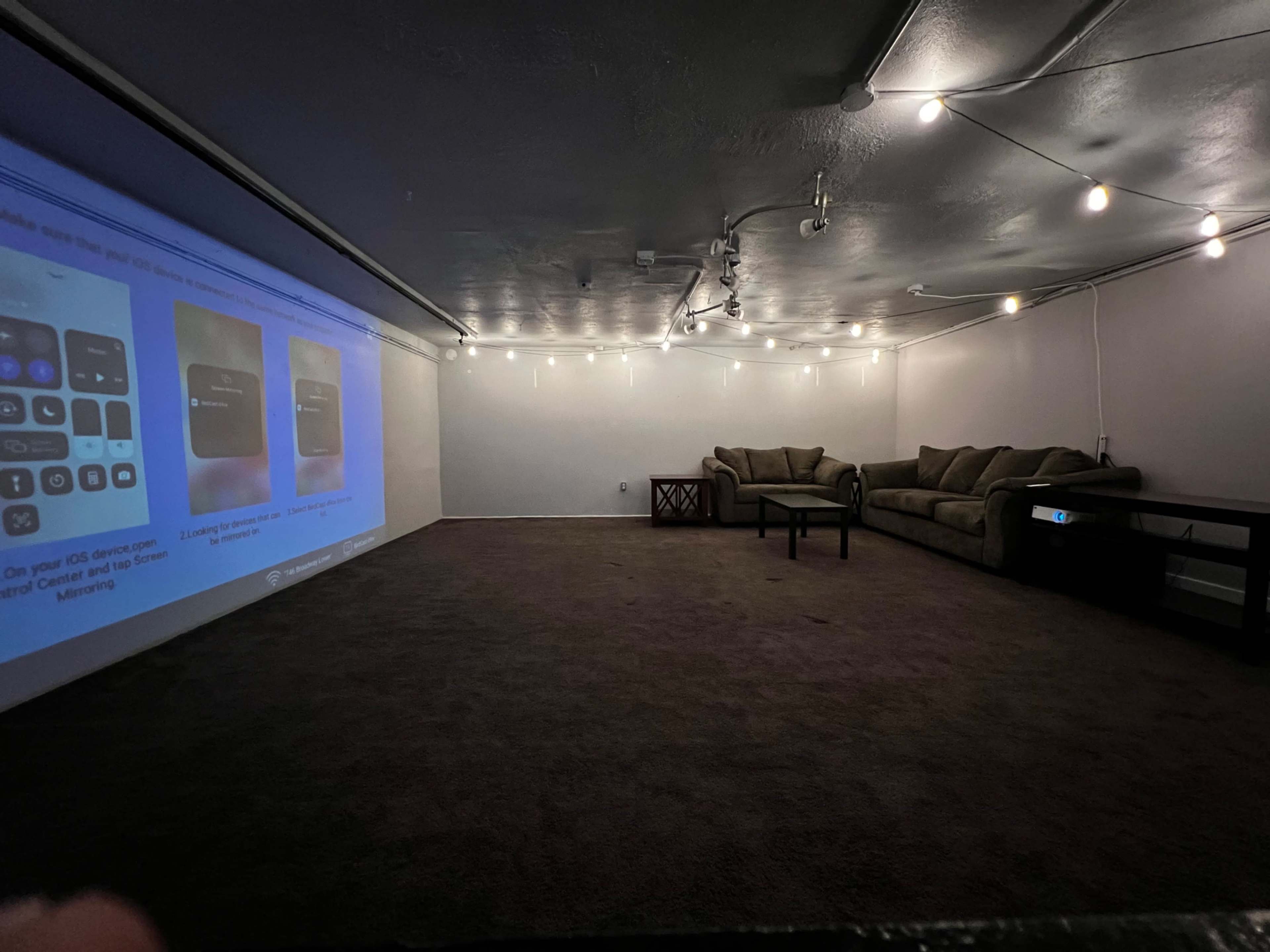 The image shows a sparsely furnished room with a projector displaying content on one wall, a sofa set, a coffee table, and string lights overhead.