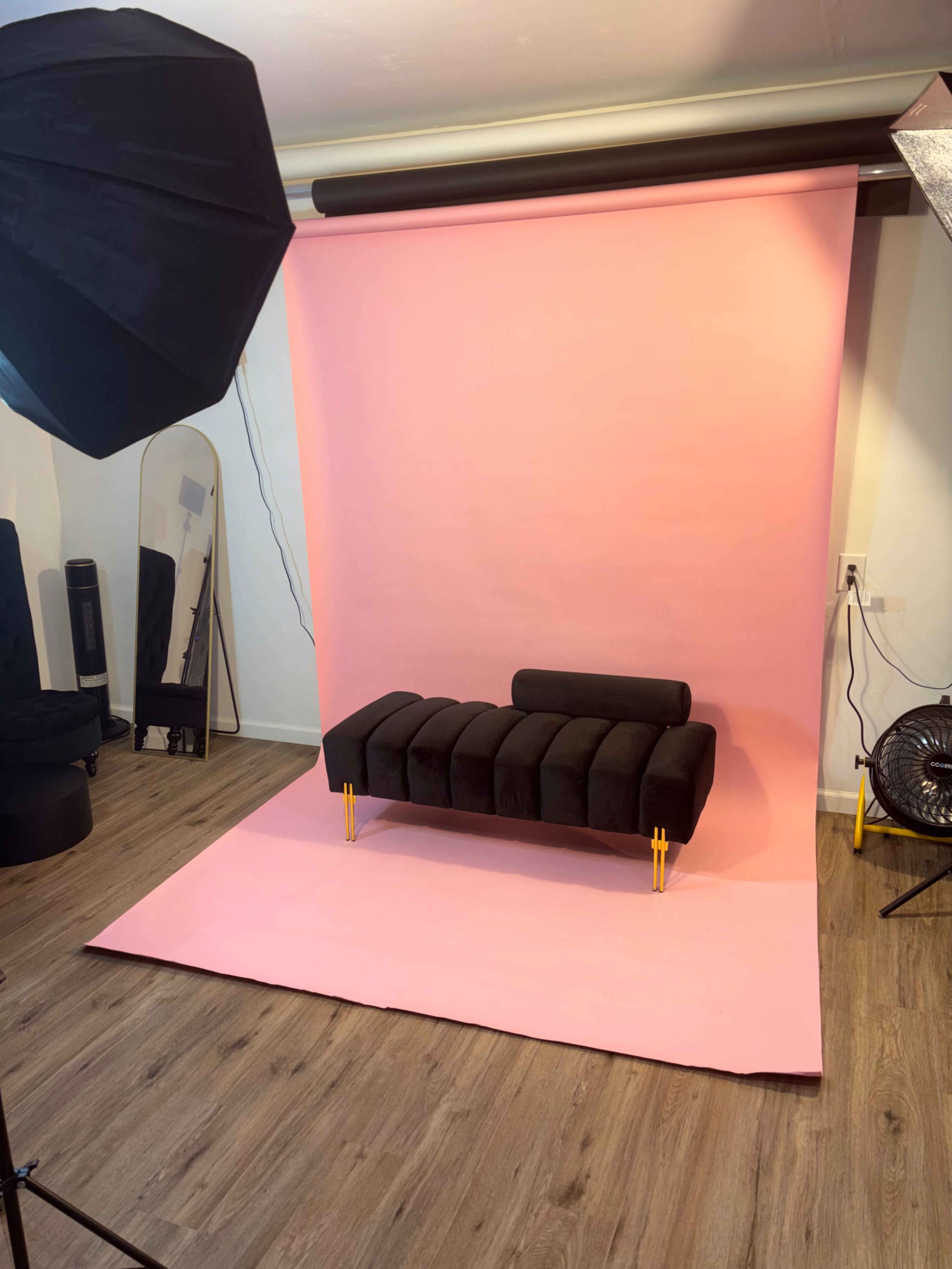 The image shows a photography studio with a black sofa positioned in front of a pink backdrop, accompanied by studio lighting equipment.