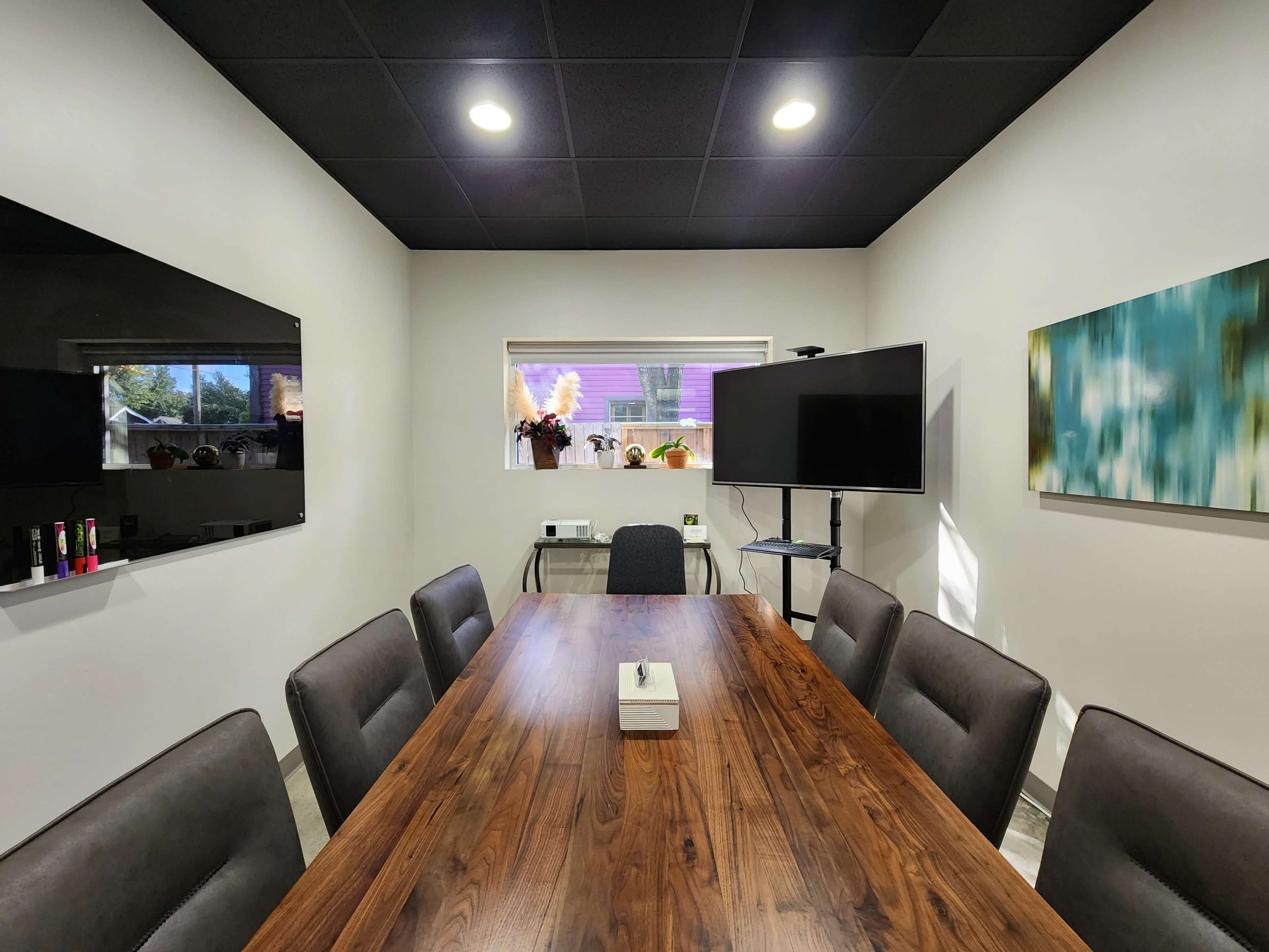 A conference room features a long wooden table surrounded by eight chairs, with two televisions mounted on the walls and a window offering natural light.