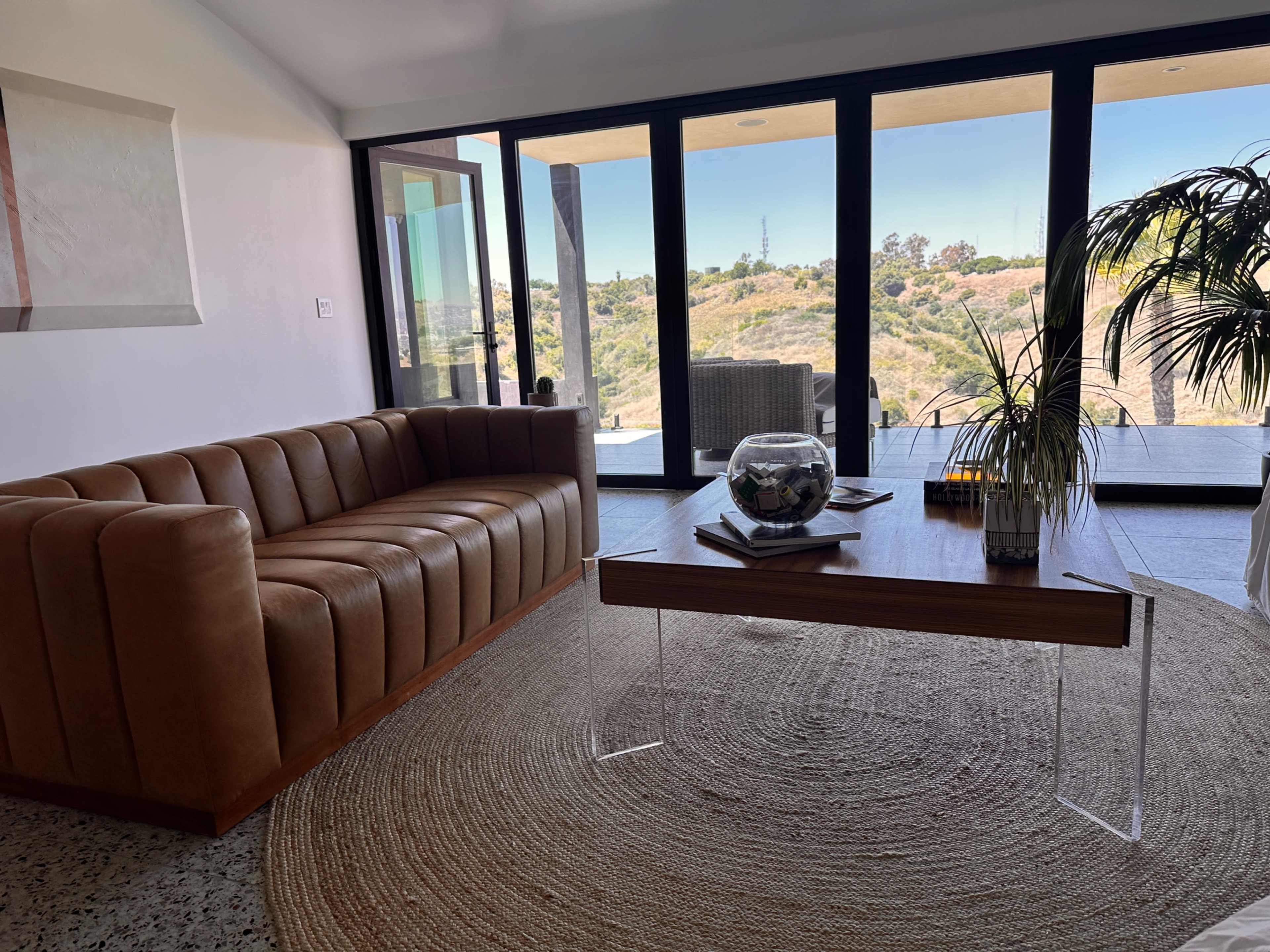 The image shows a modern living room with a brown leather sofa, a wooden table, and large glass doors leading to a view of the outdoor landscape.