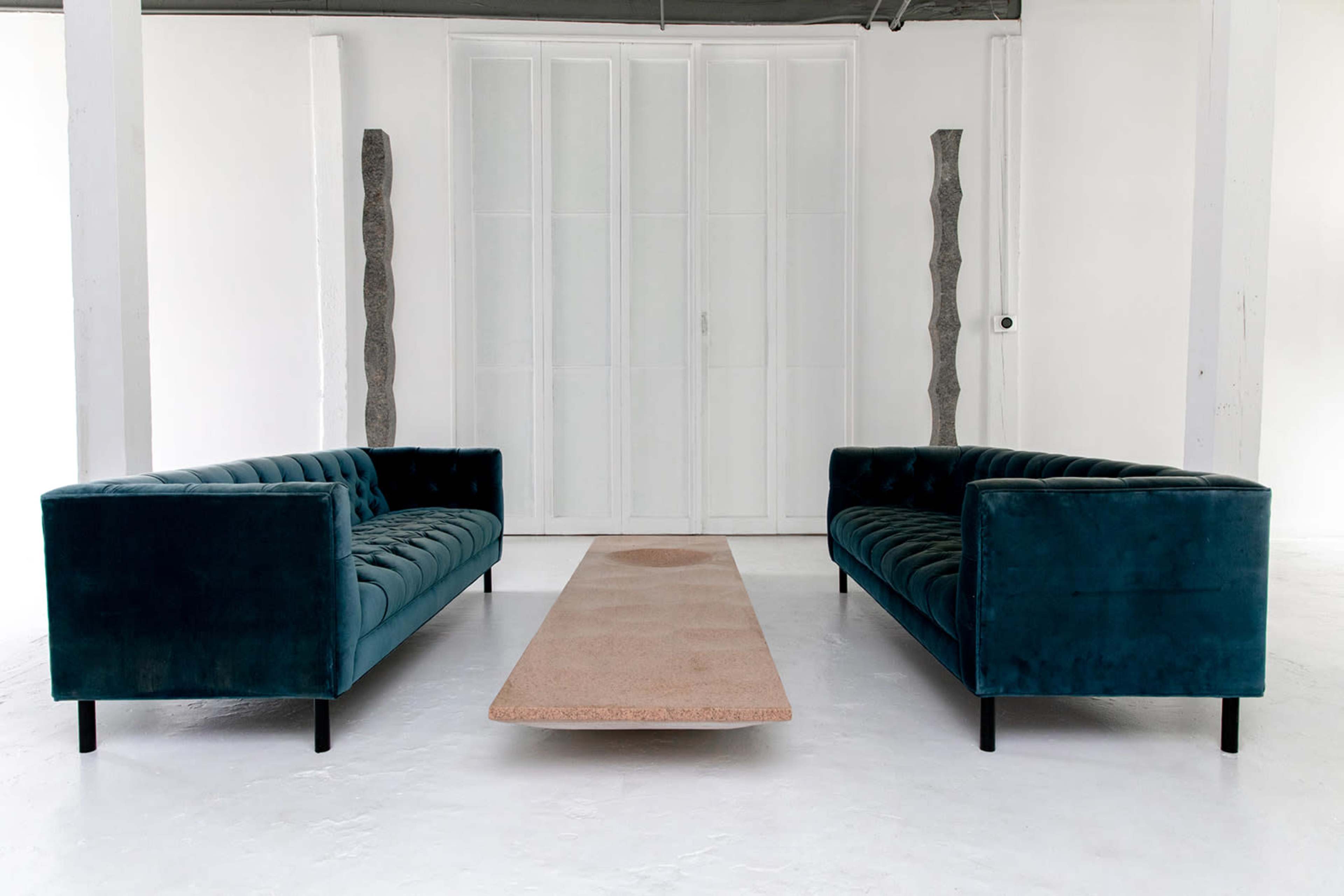 The image features two teal velvet sofas facing each other across a rectangular stone coffee table in a minimalist white room.